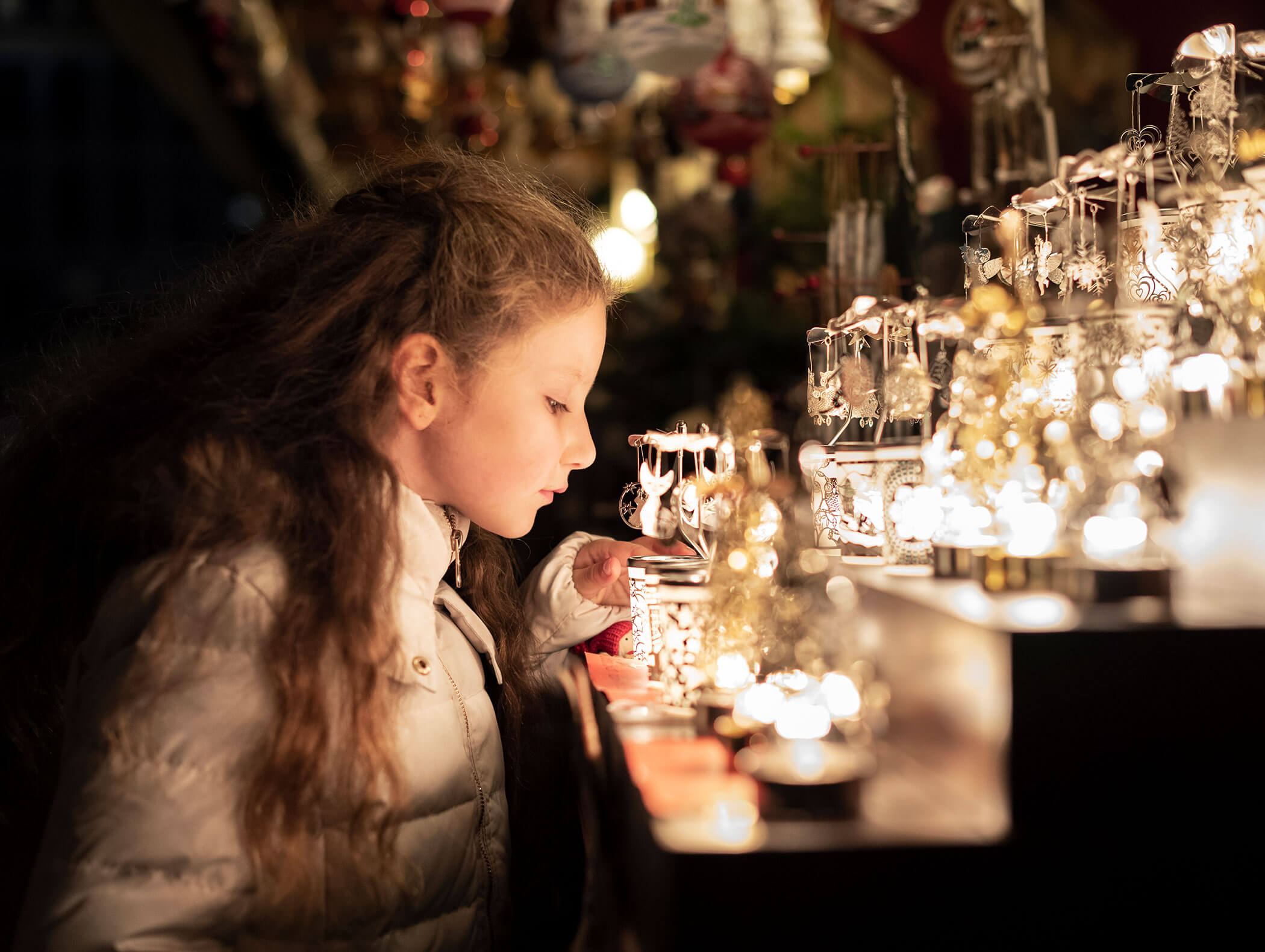 A young girl in a white coat looks closely at glowing ornaments on display at a festive market stall. - Hotel Zur Sonne