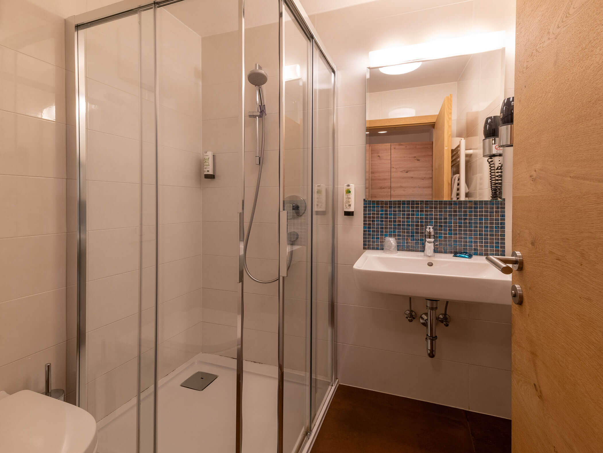 Bagno moderno con box doccia in vetro, lavabo bianco, specchio, alzatina in piastrelle blu e porta in legno. - Hotel Zur Sonne