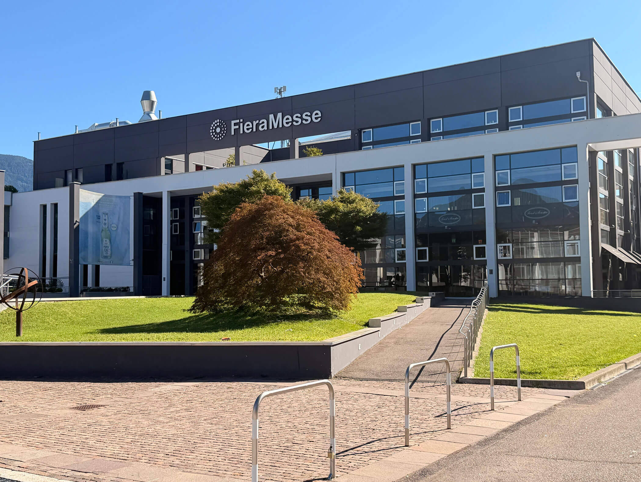 Modernes Gebäude mit der Aufschrift „FieraMesse“, Glasfenstern, einer grünen Rasenfläche und einem Baum in der Mitte davor. - Hotel Zur Sonne