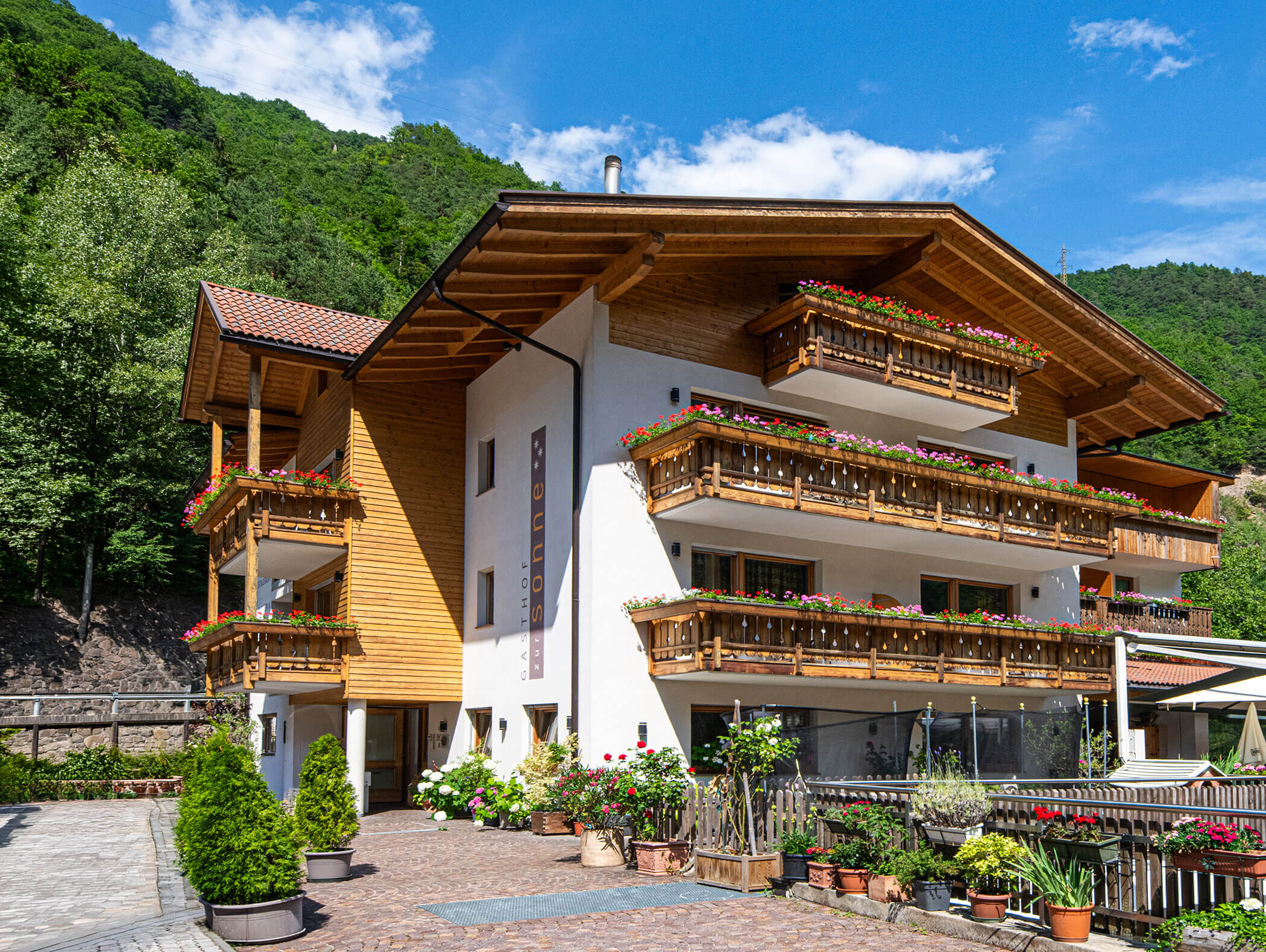 Alpine-style hotel with wooden balconies and flower boxes, surrounded by greenery on a sunny day. - Hotel Zur Sonne