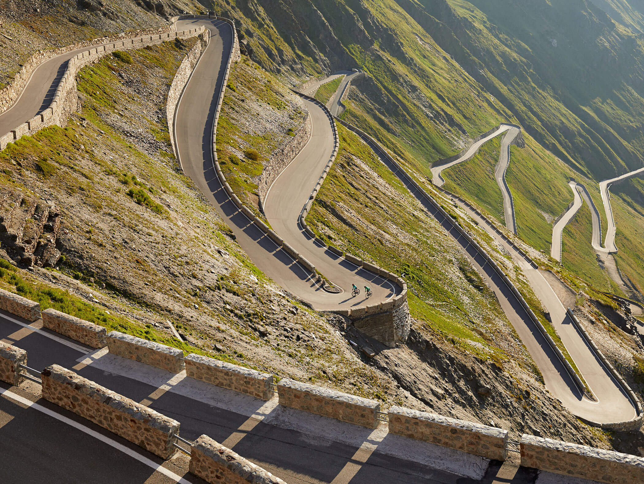 A winding mountain road with many sharp hairpin turns on a steep, grassy slope. - Hotel Zur Sonne