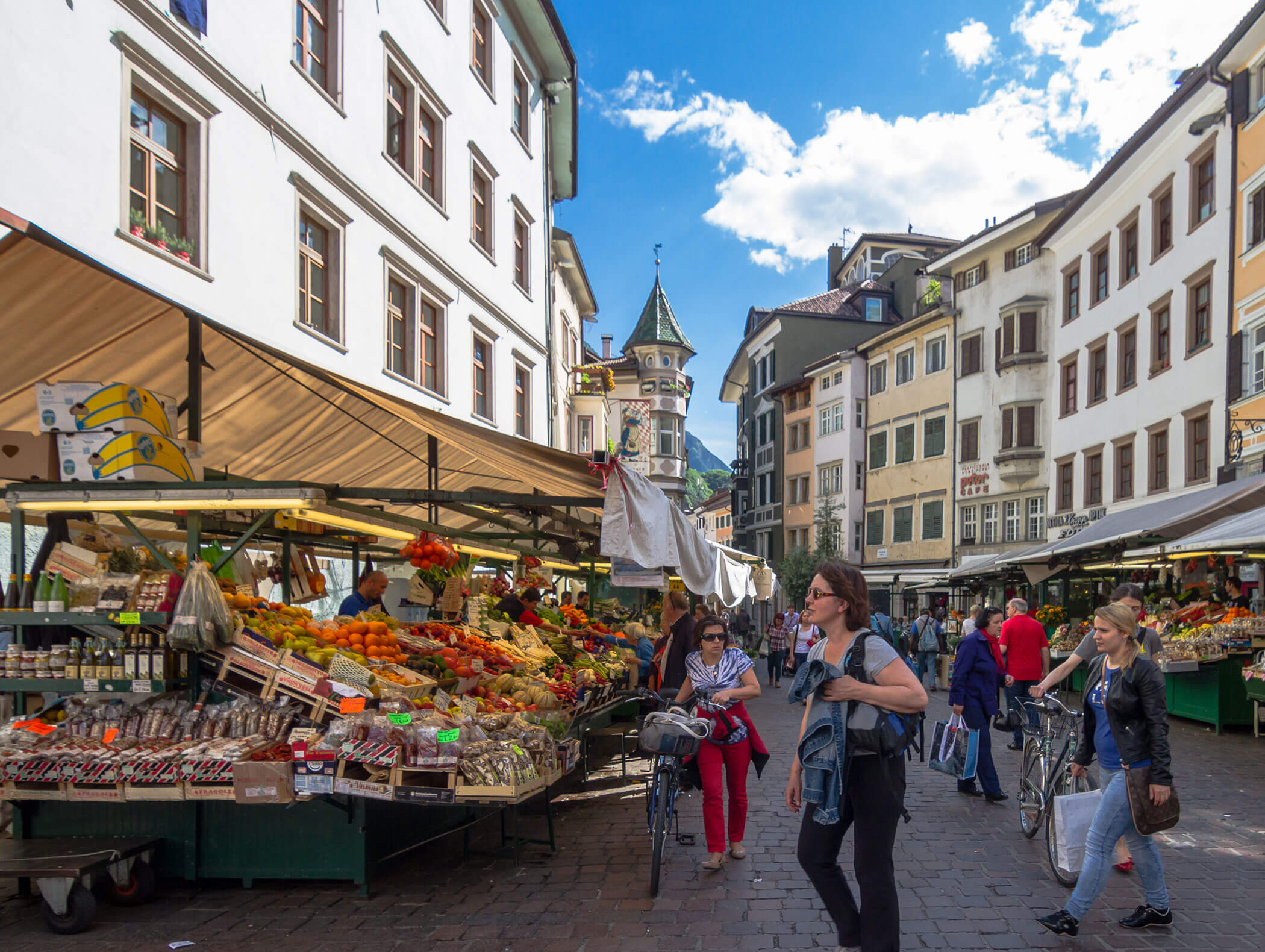 People walk by colorful market stalls with fruits and vegetables on a lively city street under a blue sky. - Hotel Zur Sonne