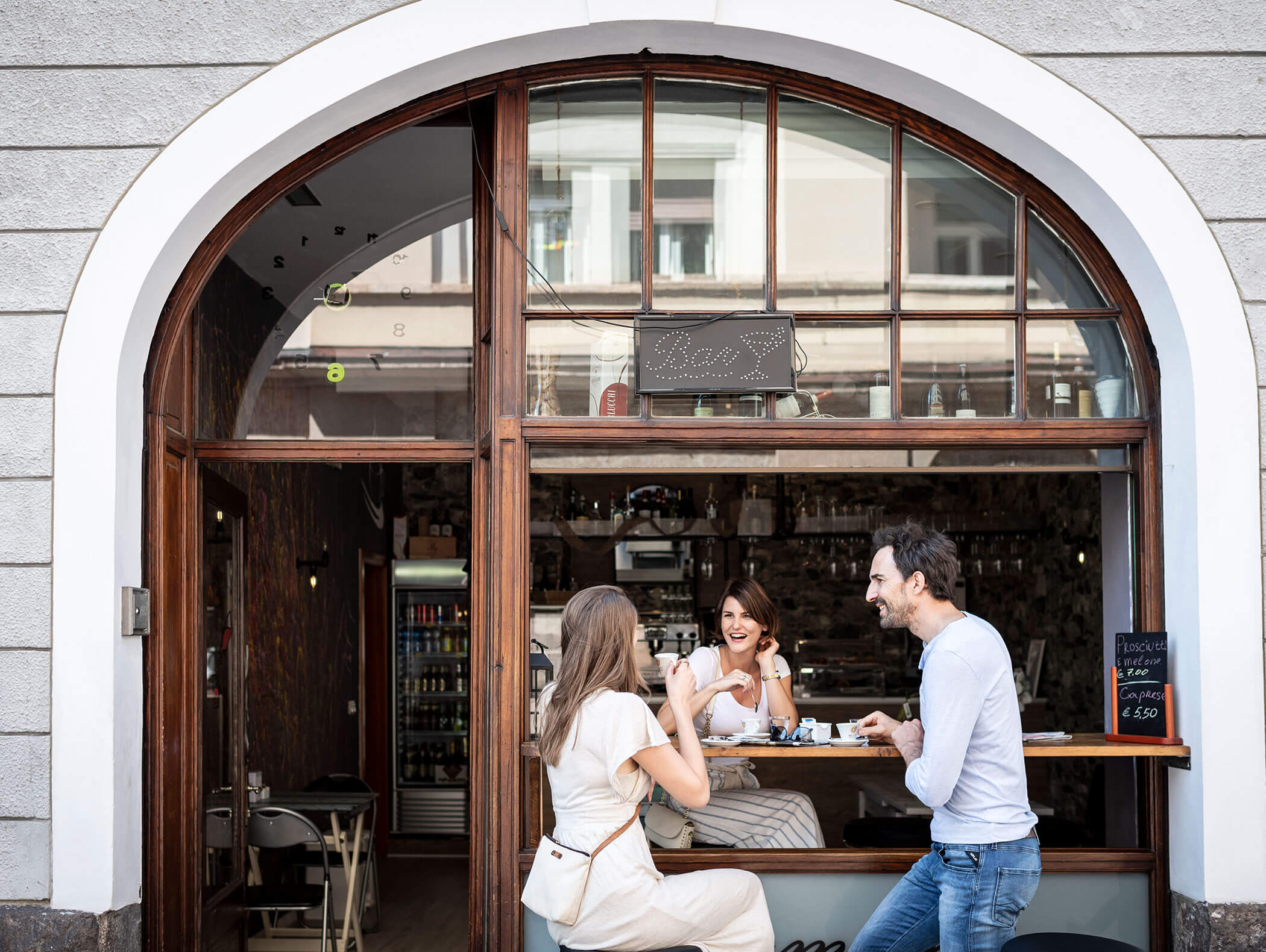 Three people enjoy drinks and conversation outside a café with a large arched window. - Hotel Zur Sonne