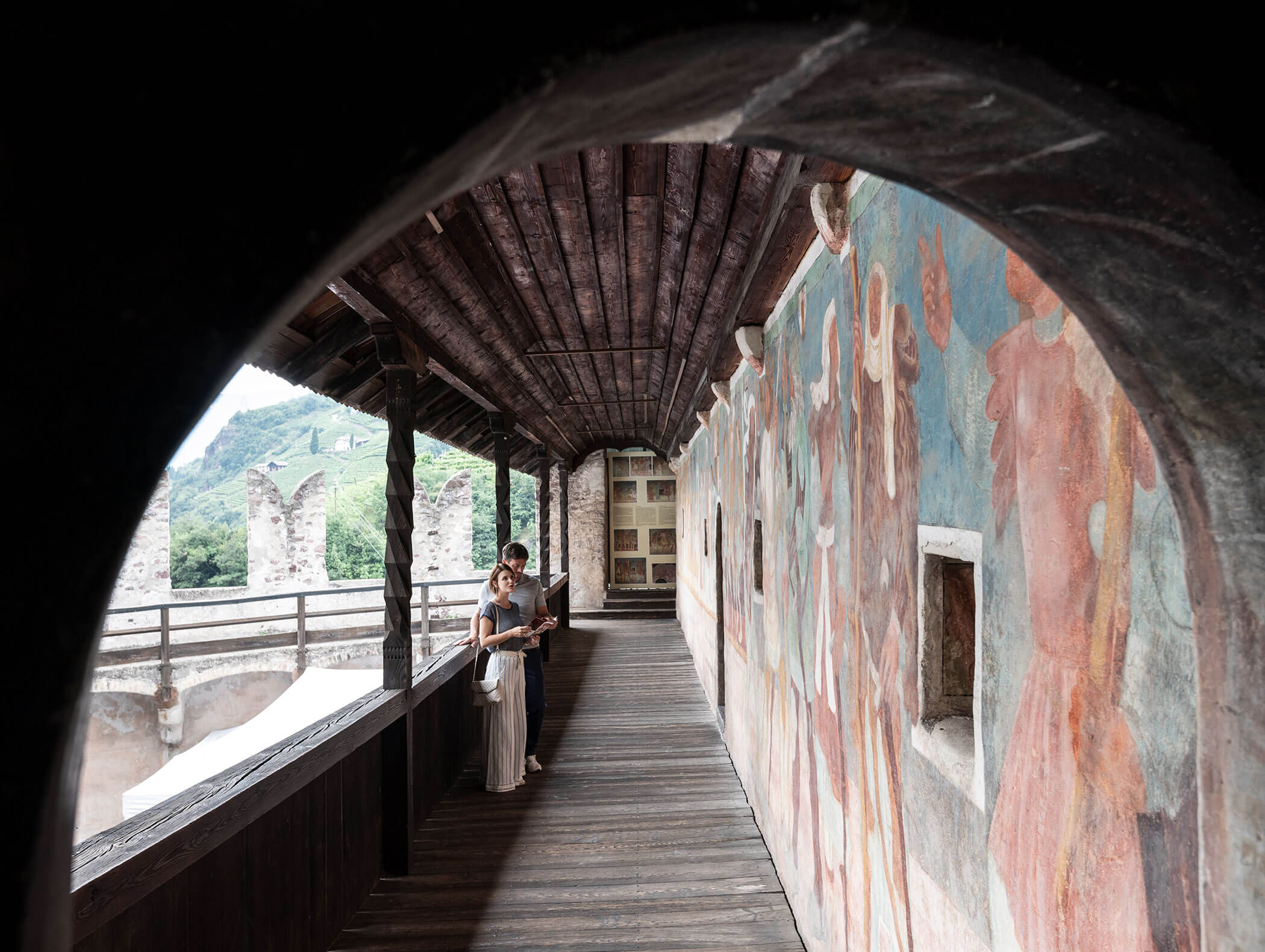 A person stands on a wooden balcony with colorful murals and mountain scenery visible through an archway. - Hotel Zur Sonne