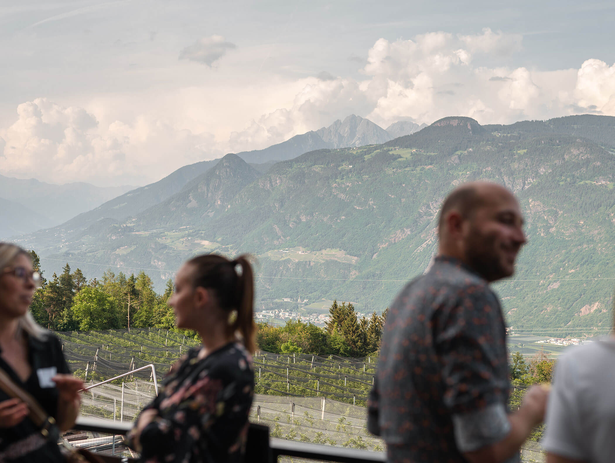 Three people converse outdoors with mountains and cloudy sky in the background. - Hotel Zur Sonne