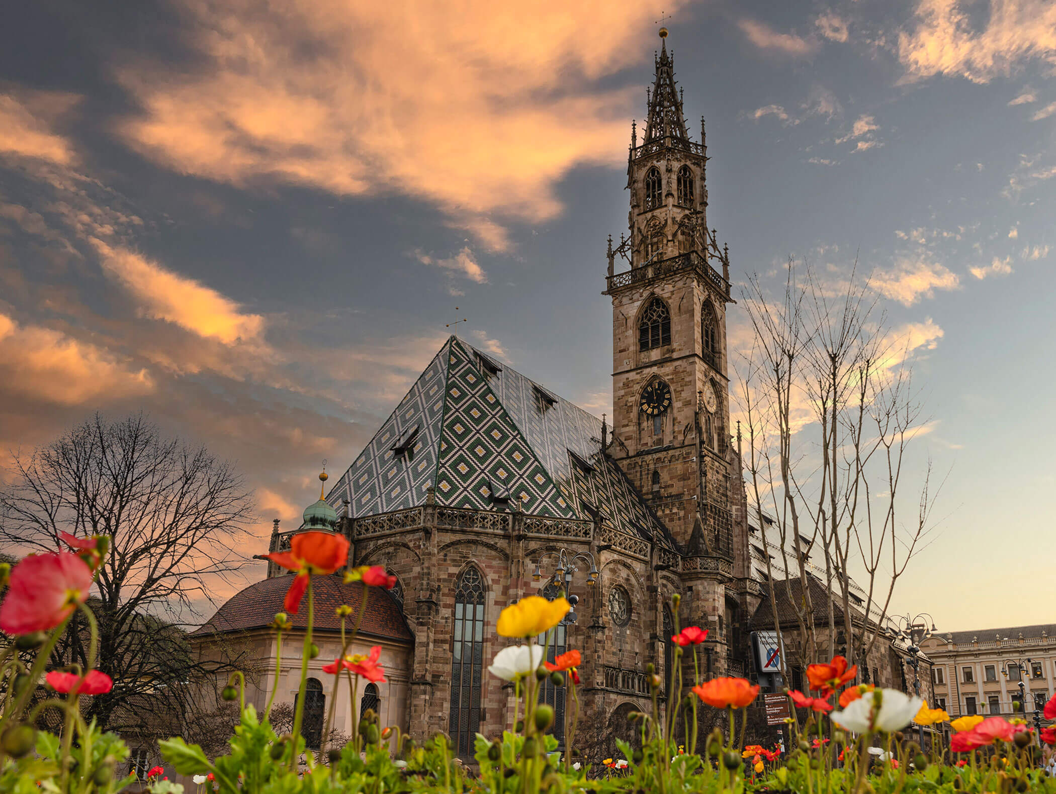 Gothic church with patterned roof and tall tower at sunset, with colorful flowers in the foreground. - Hotel Zur Sonne