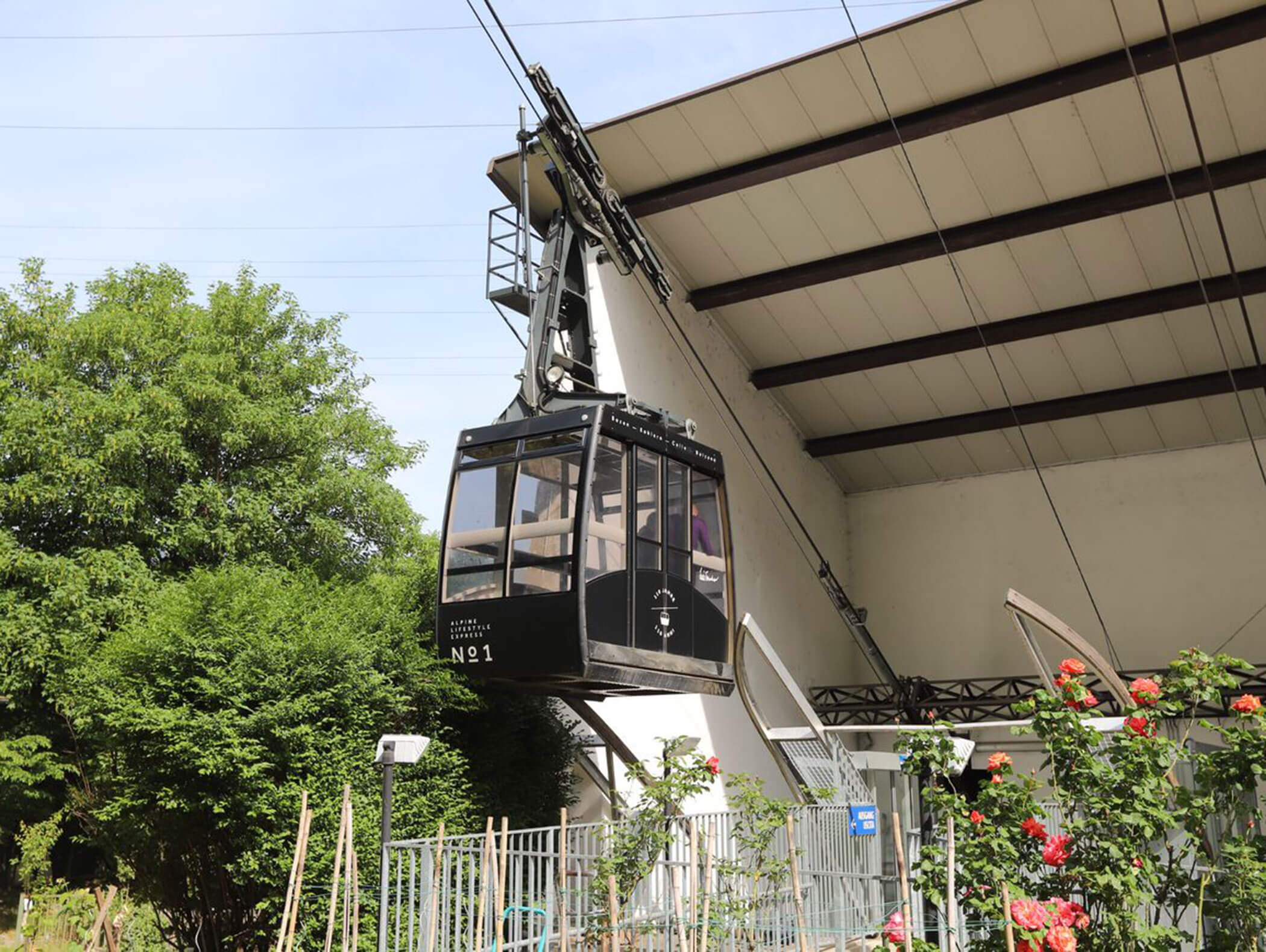 A black cable car leaves a station, surrounded by greenery and flowers on a sunny day. - Hotel Zur Sonne