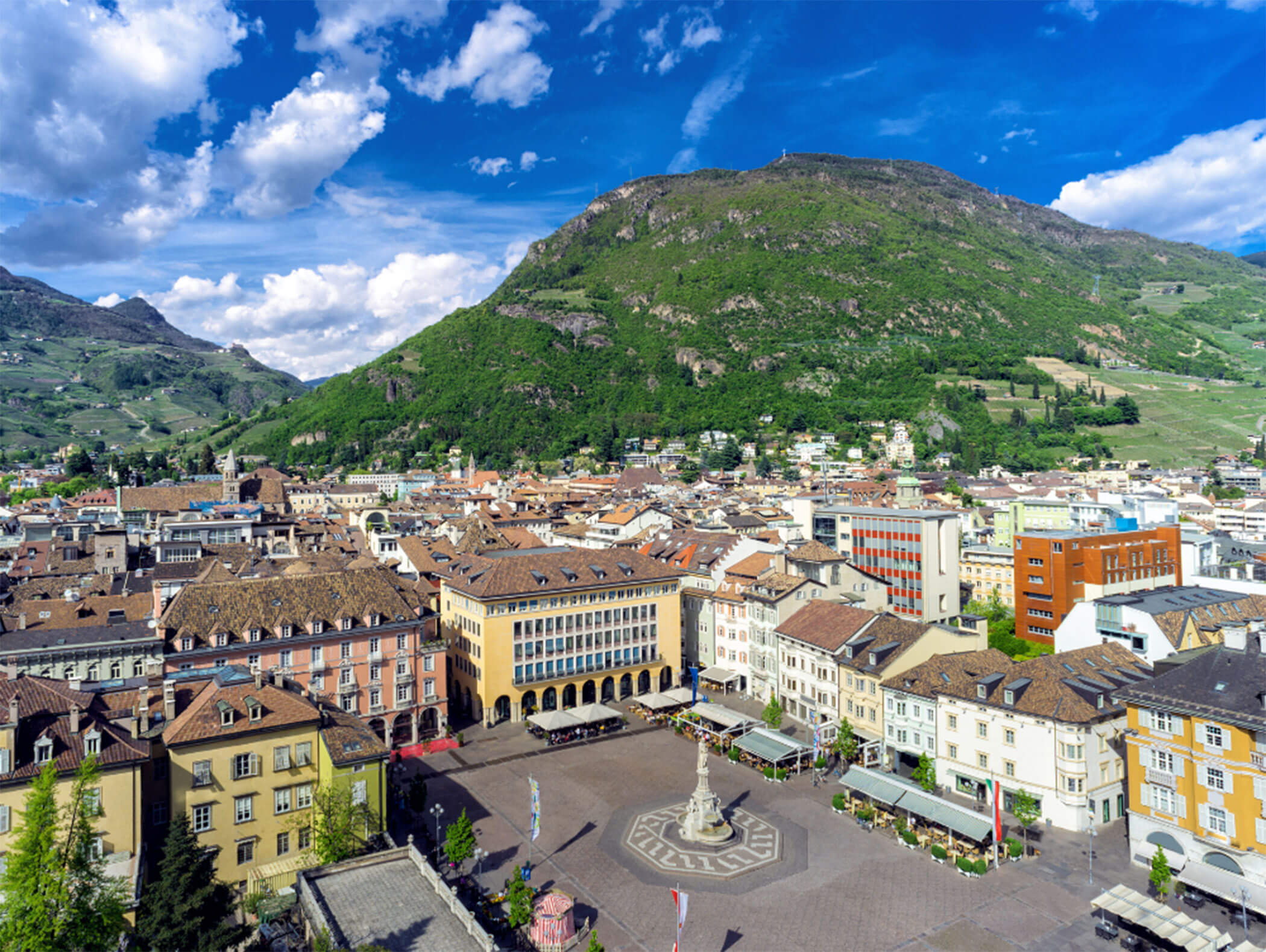 Town square with a fountain, surrounded by colorful buildings and a green hill in the background under blue sky. - Hotel Zur Sonne
