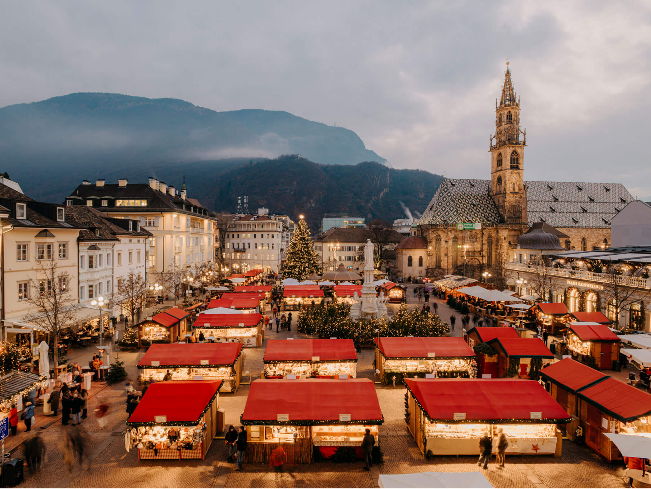 Christmas market with red-roofed stalls, a large tree, and a church, set against a mountain backdrop at dusk. - Hotel Zur Sonne