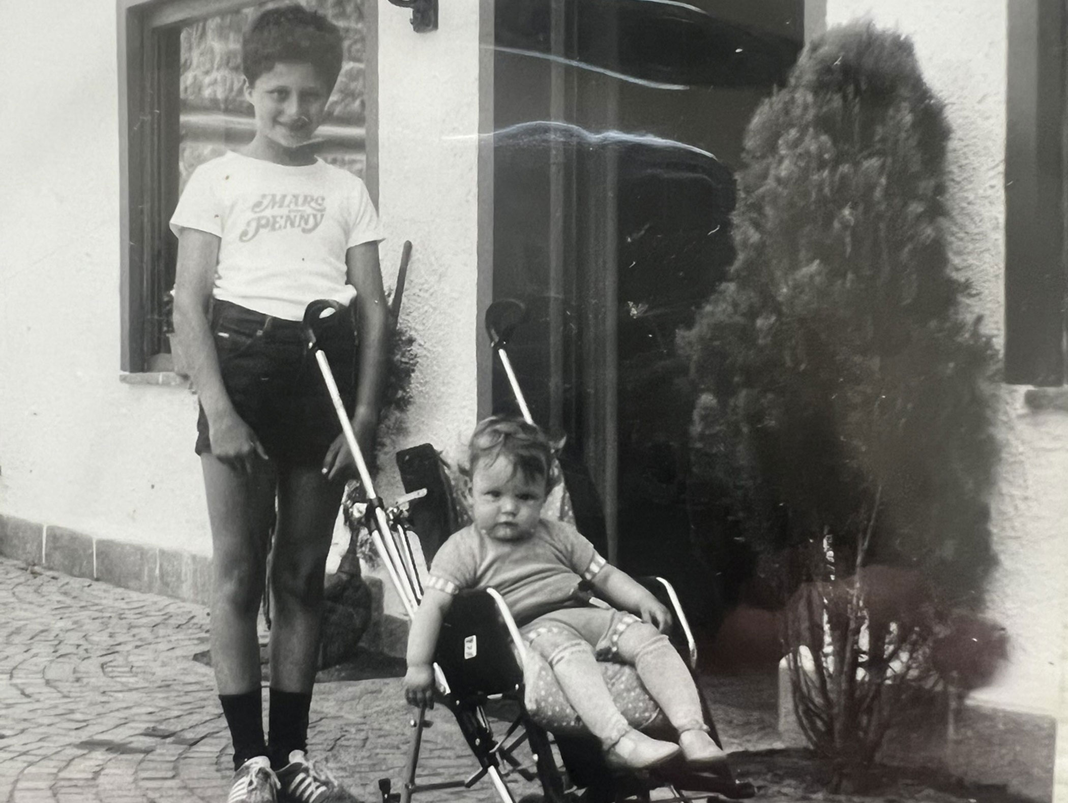 Ein lächelnder Junge steht neben einem Kleinkind, das in einem Kinderwagen vor einem Gebäude sitzt – Schwarz-Weiß-Foto. - Hotel Zur Sonne