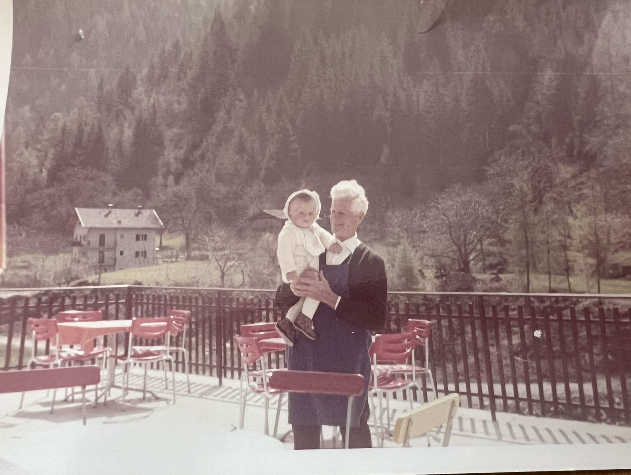 Ein älterer Mann hält ein Baby auf einer Terrasse mit roten Stühlen, Bäumen und einem Haus im Hintergrund im Arm. - Hotel Zur Sonne