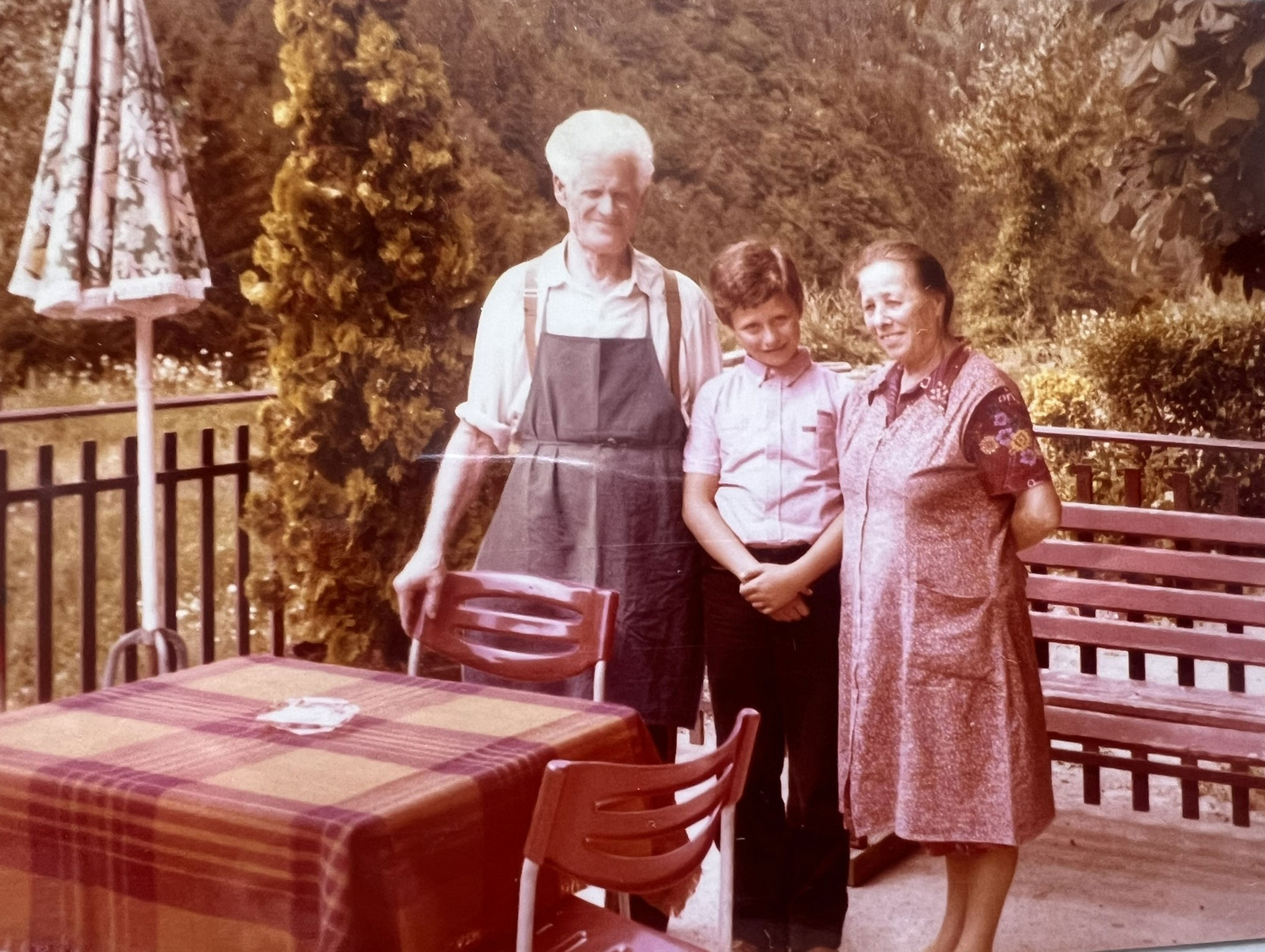 Ein älterer Mann, ein Junge und eine Frau stehen gemeinsam im Freien an einem Tisch mit einer rot karierten Tischdecke. - Hotel Zur Sonne