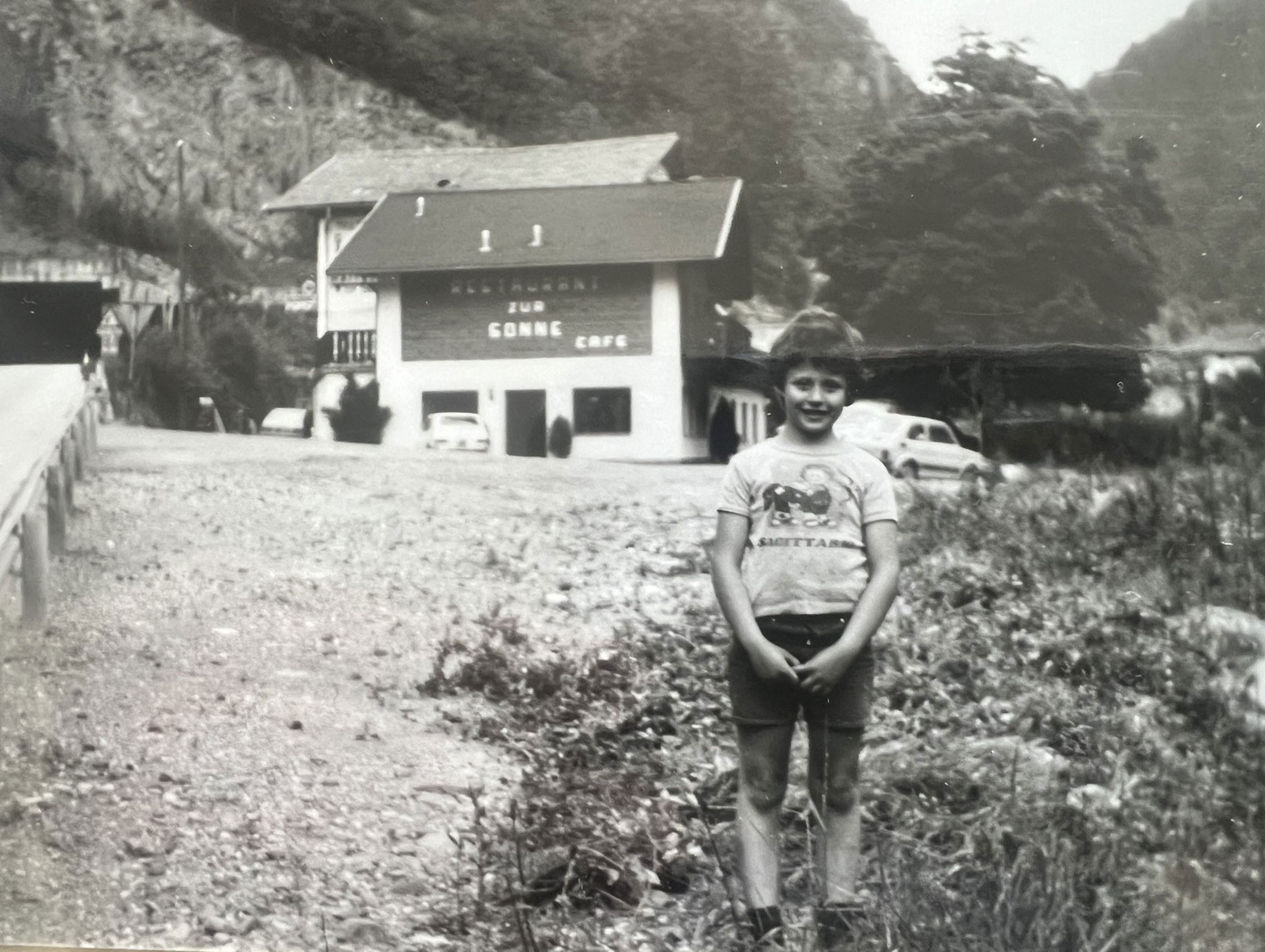 Ein lächelnder Junge steht auf einem Feldweg, im Hintergrund sind ein Gebäude, Autos und Berge zu sehen. - Hotel Zur Sonne