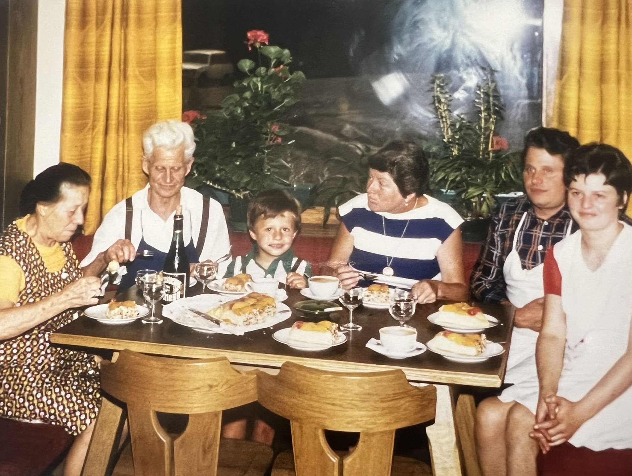 Sechs Menschen unterschiedlichen Alters sitzen an einem Tisch mit Essen und Getränken und genießen gemeinsam eine Mahlzeit in einem gemütlichen Raum. - Hotel Zur Sonne