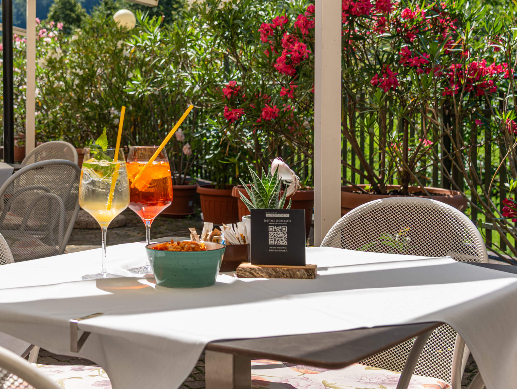 Two cocktails and a bowl of snacks on a sunny outdoor café table with flowers in the background. - Hotel Zur Sonne