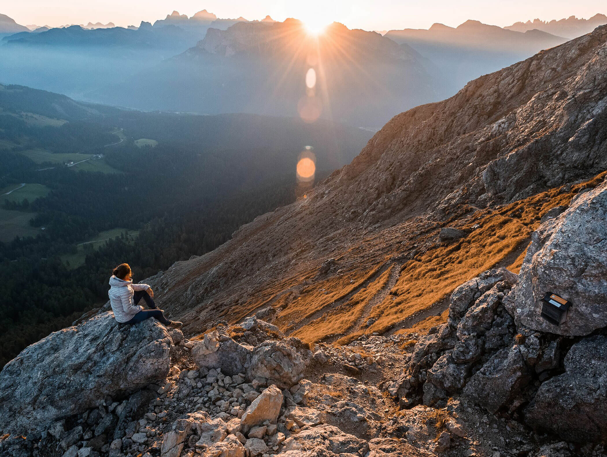 Eine Person sitzt bei Sonnenaufgang auf einem felsigen Berghang und blickt auf ein nebliges Tal und ferne Berggipfel. - Hotel Zur Sonne