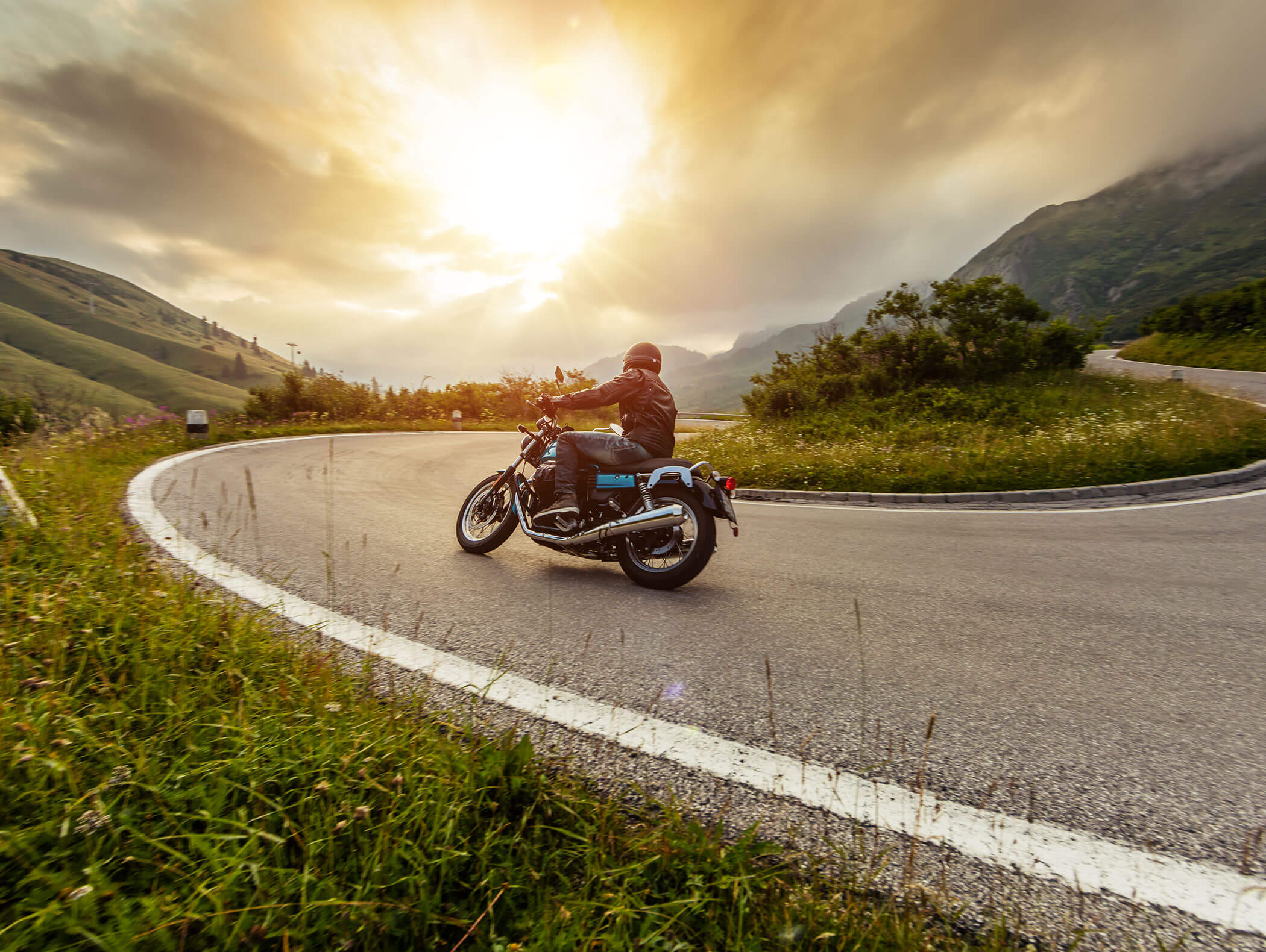 Eine Person fährt bei Sonnenuntergang mit dem Motorrad um eine Kurve auf einer malerischen Bergstraße, umgeben von üppigem Grün. - Hotel Zur Sonne