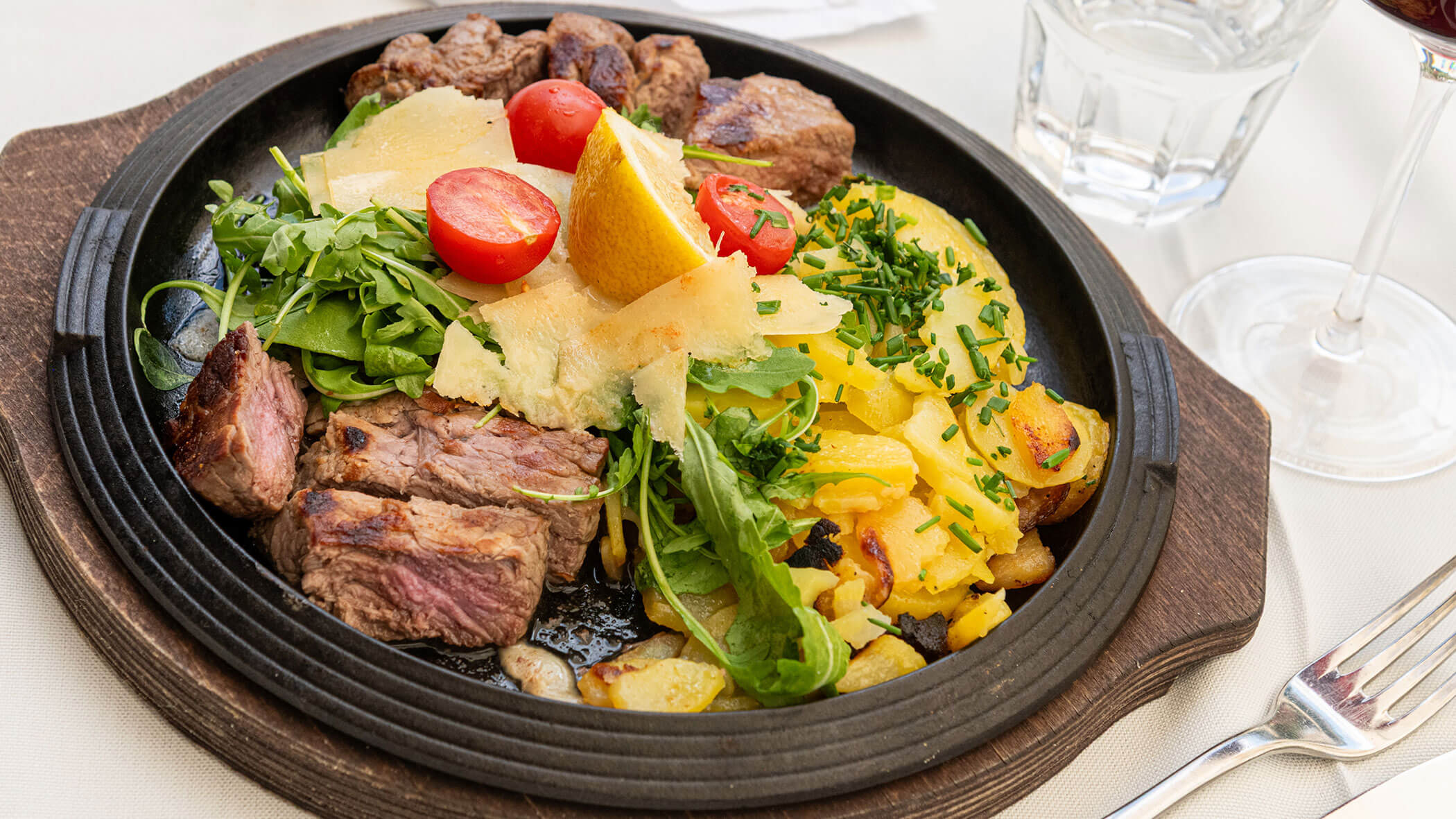 Steak in Scheiben, Bratkartoffeln, Rucola, Kirschtomaten und Parmesan auf einem schwarzen Teller, dazu ein Glas Wein. - Hotel Zur Sonne