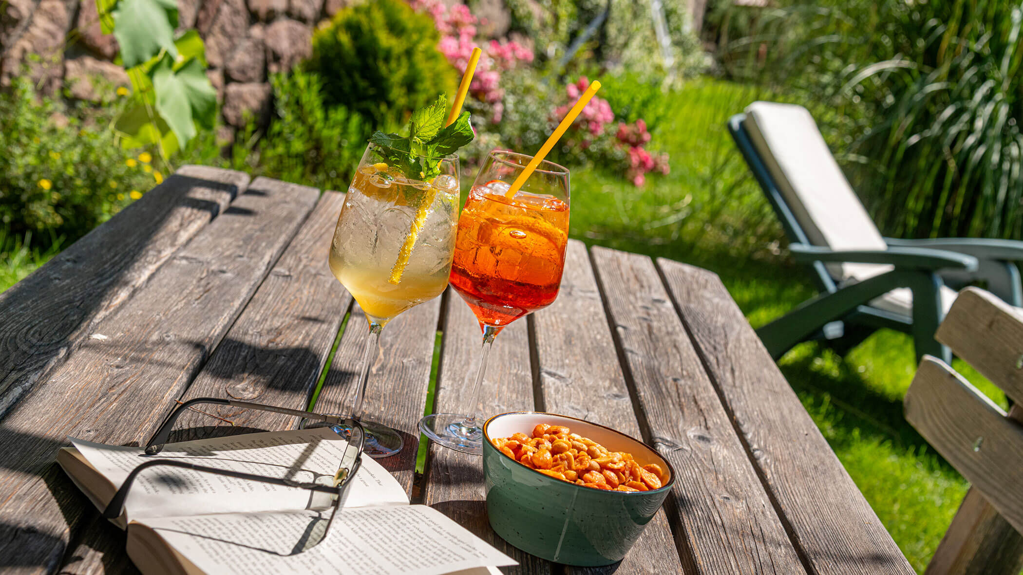 Zwei Cocktails, eine Schale mit Knabbereien, ein Buch und Gläser auf einem Holztisch in einem sonnigen Garten. - Hotel Zur Sonne