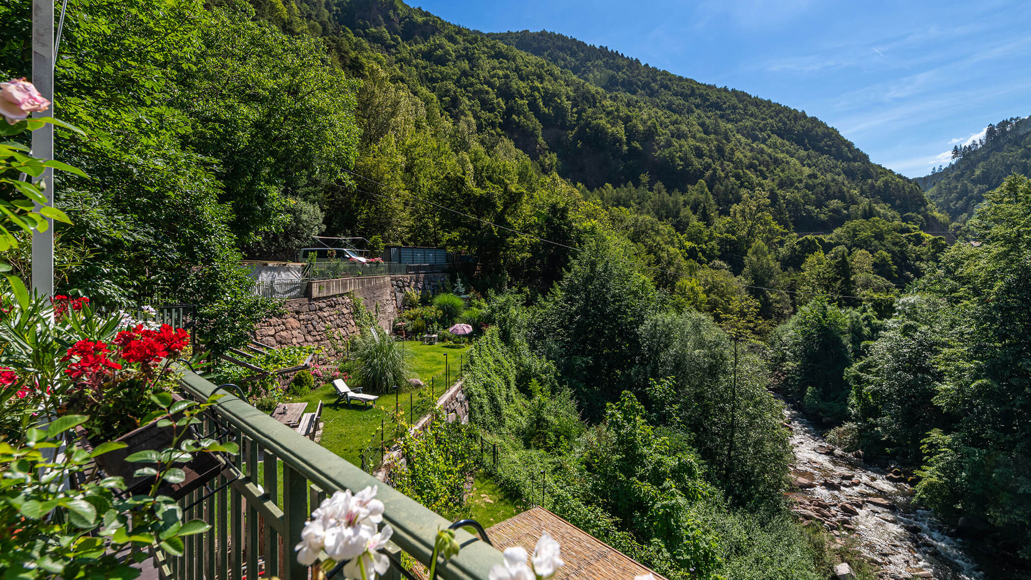 Der mit Blumen geschmückte Balkon bietet unter strahlend blauem Himmel einen Blick auf ein üppig grünes Tal, einen Fluss und bewaldete Hügel. - Hotel Zur Sonne
