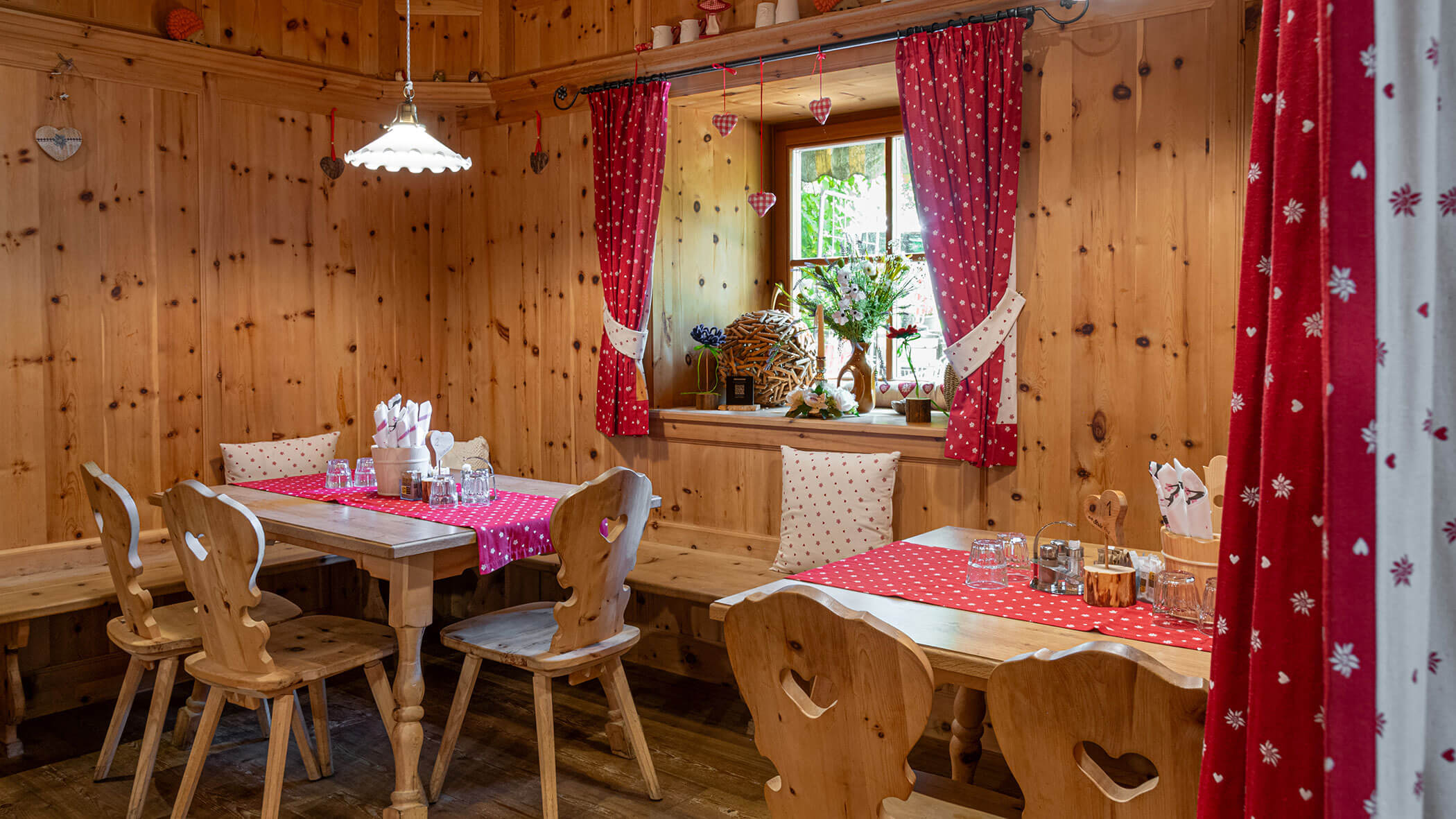 Gemütliches Esszimmer aus Holz mit roten Vorhängen, zwei Tischen, Stühlen und einem kleinen, mit Blumen geschmückten Fenster. - Hotel Zur Sonne