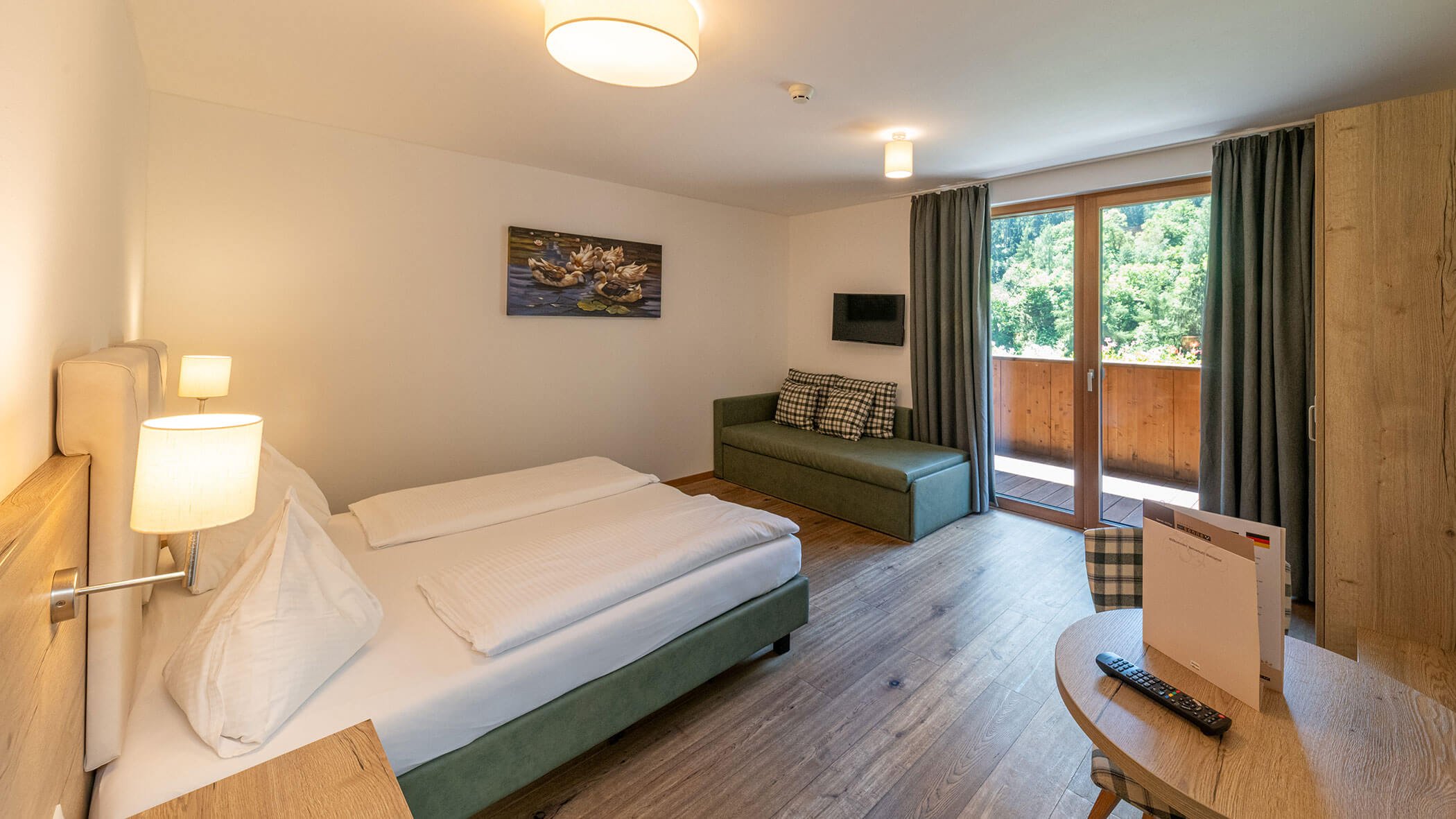 Helles Hotelzimmer mit einem Doppelbett, einem Sofa, einem Fernseher, einem Balkon, einem Holzboden und Blick ins Grüne durch die Glastüren. - Hotel Zur Sonne