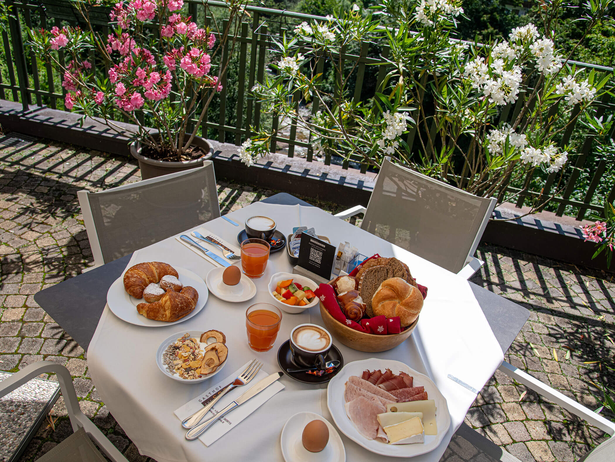 Tavolo da colazione all’aperto con cornetti, succo di frutta, salumi, uova, frutta e fiori sullo sfondo. - Hotel Zur Sonne
