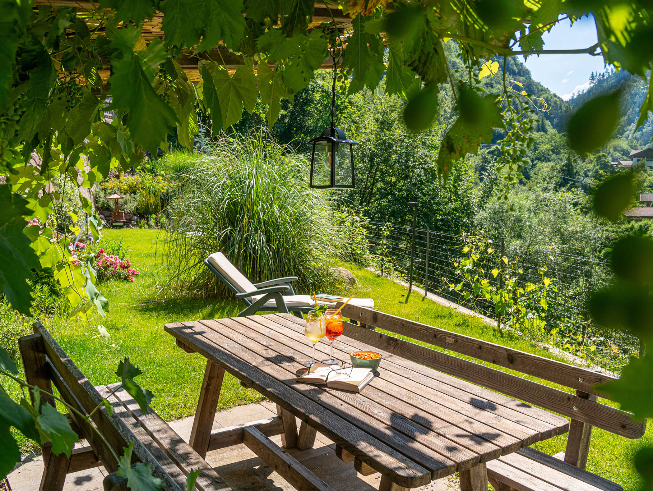 Tavolo da picnic in legno con bevande e libri, immerso nel verde, in un giardino soleggiato con una sdraio e le colline sullo sfondo. - Hotel Zur Sonne