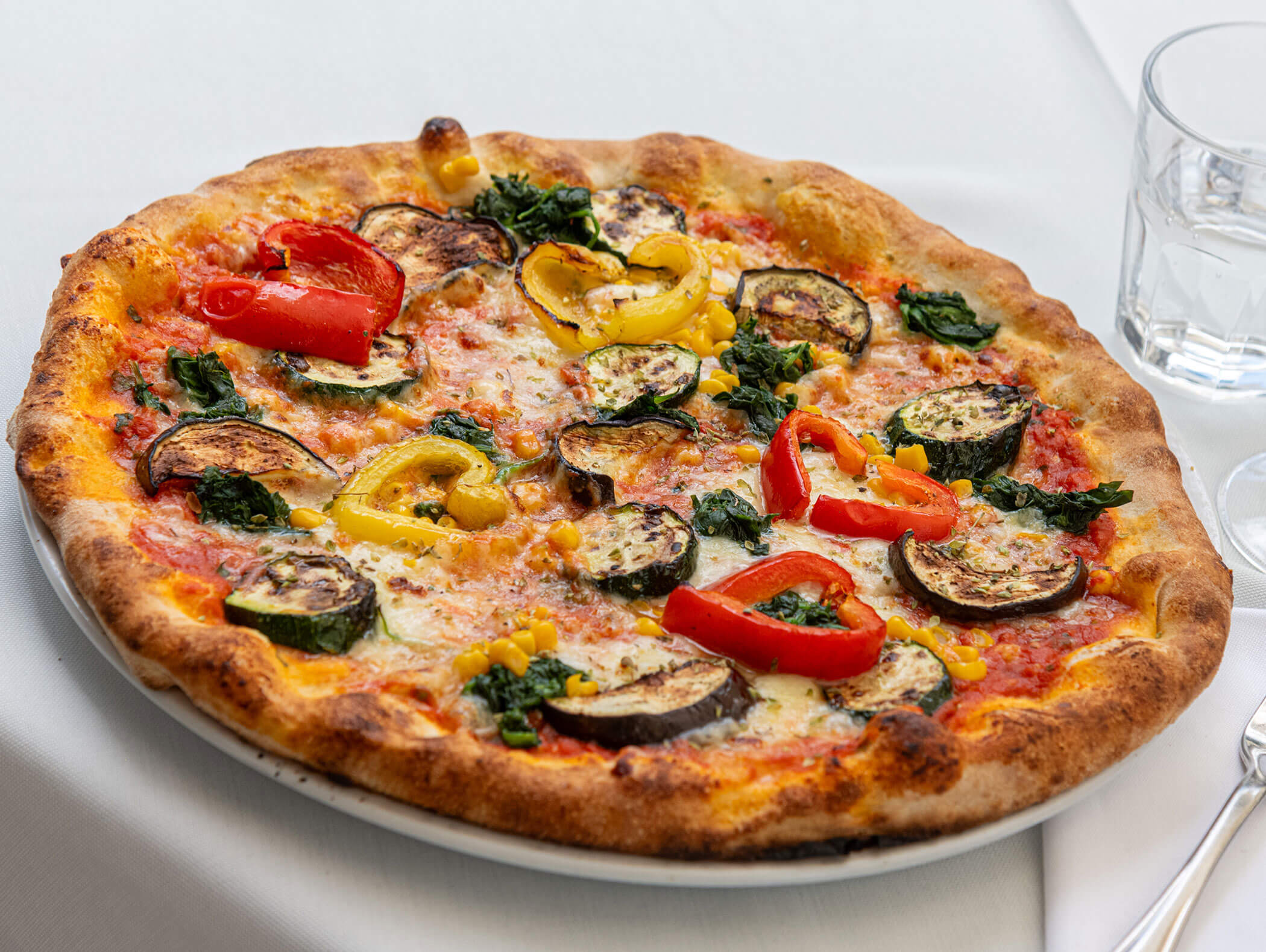 A vegetable pizza with zucchini, bell peppers, spinach, and corn on a plate next to a glass of water. - Hotel Zur Sonne