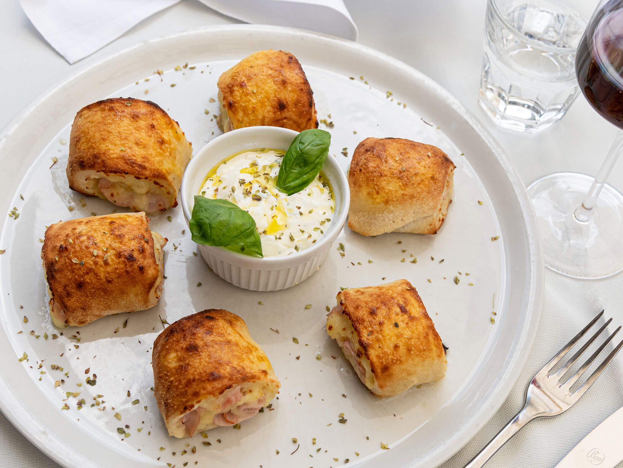 Sei panini cotti al forno disposti attorno a una ciotola di salsa, guarniti con basilico. Accanto ci sono una forchetta, un bicchiere d’acqua e un bicchiere da vino. - Hotel Zur Sonne
