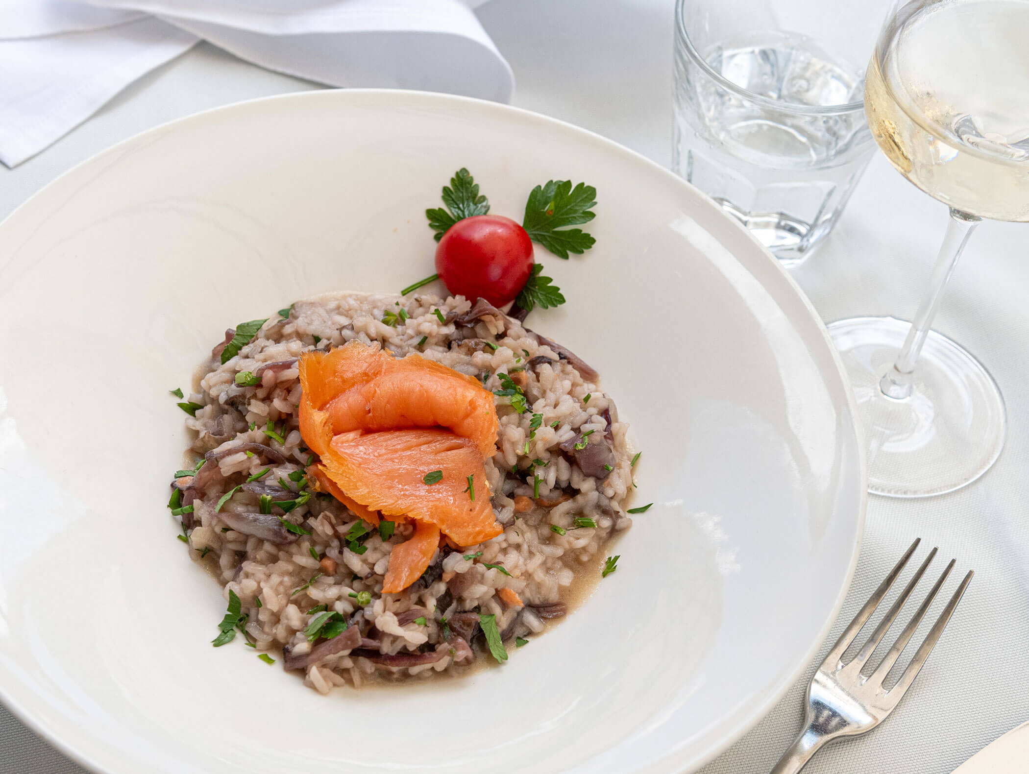 Piatto di risotto guarnito con salmone affumicato, prezzemolo e pomodoro, accanto a un bicchiere di vino bianco e un bicchiere d’acqua. - Hotel Zur Sonne