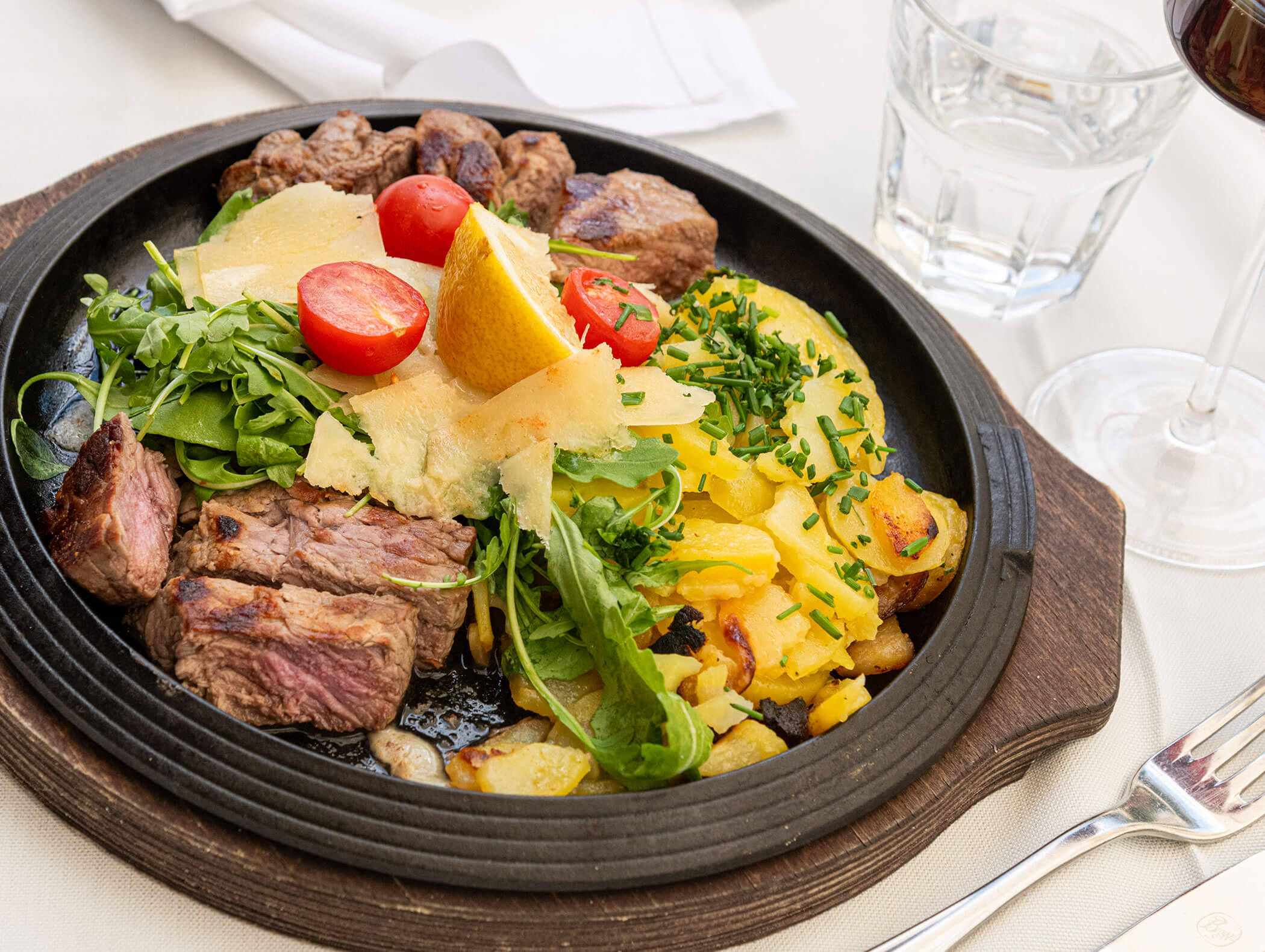Grilled steak with arugula, cherry tomatoes, cheese, roasted potatoes, lemon, and a glass of water. - Hotel Zur Sonne