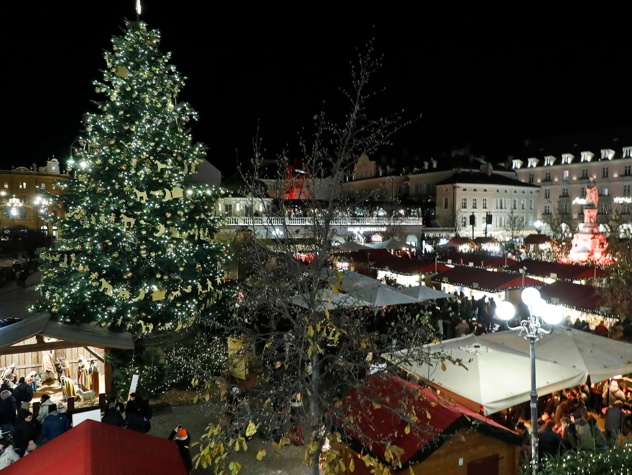 A festive Christmas market at night with a large decorated tree and crowded stalls under lights. - Hotel Zur Sonne