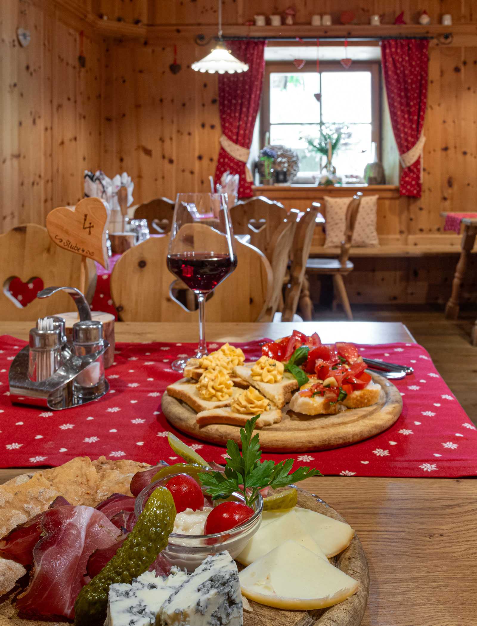 Ein Holztisch mit Käse, Aufschnitt, Brot und einem Glas Rotwein in einem gemütlichen, rustikalen Speisesaal. - Hotel Zur Sonne