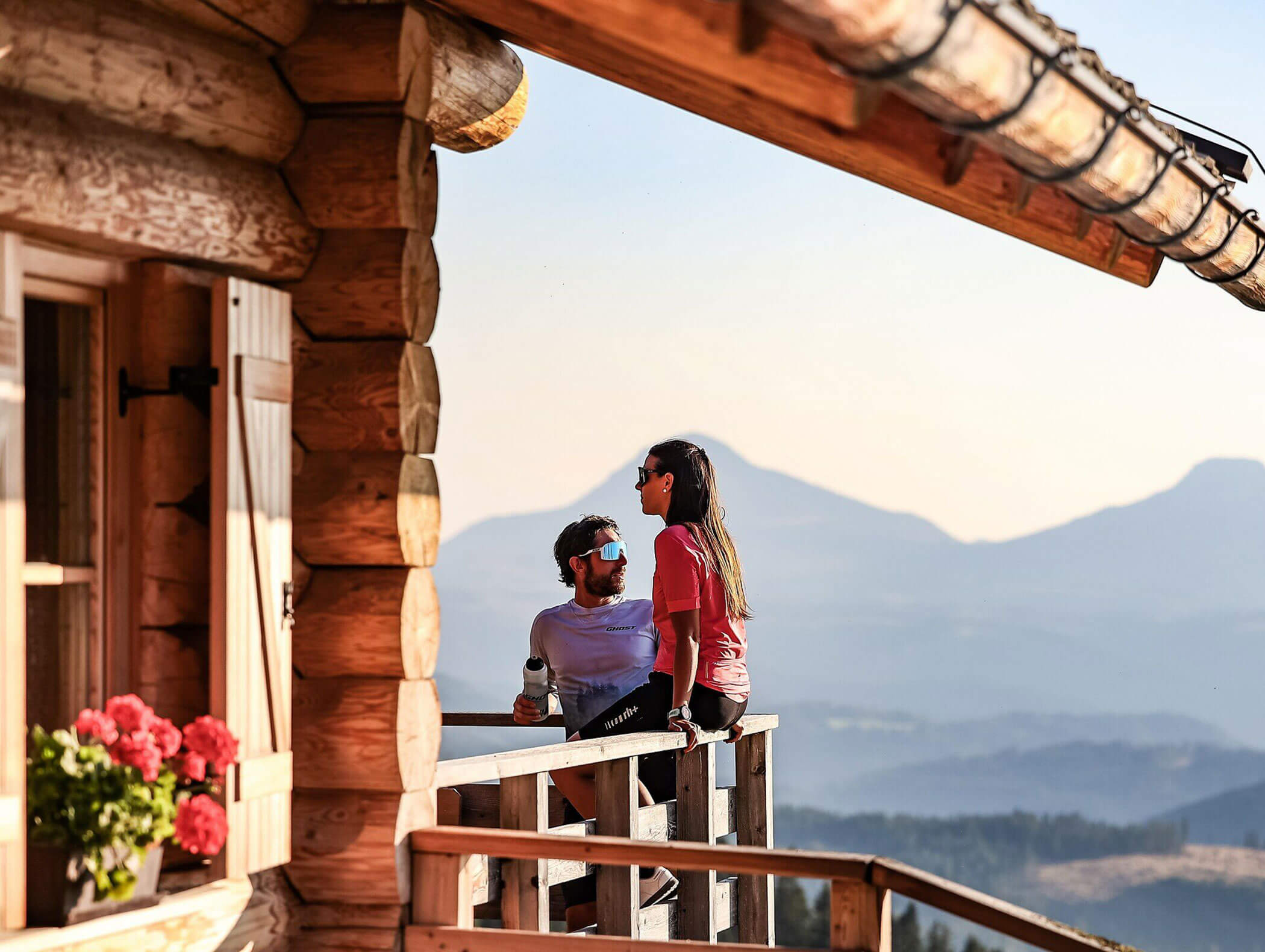 A couple relaxes on a wooden cabin balcony with mountains and flowers in the background. - Hotel Zur Sonne