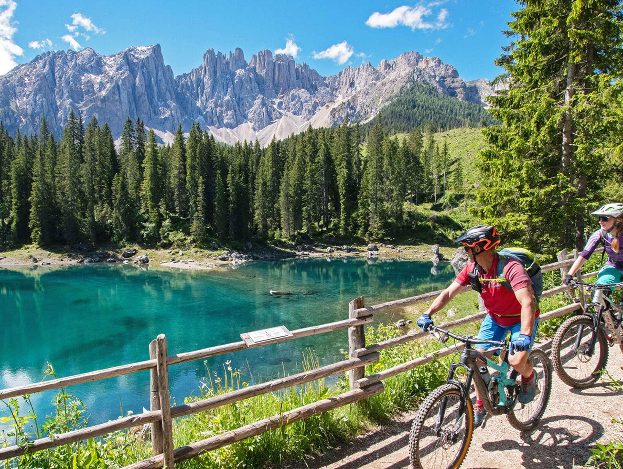 Two cyclists ride by a turquoise lake with pine trees and rocky mountains under a clear blue sky. - Hotel Zur Sonne