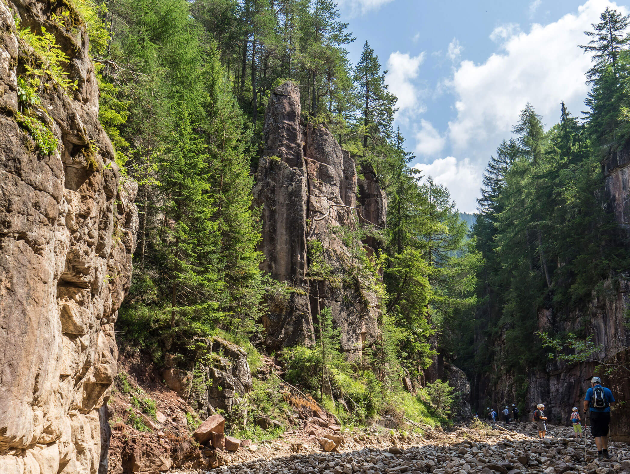 Hikers walk through a rocky canyon lined with tall trees and steep cliff walls under a partly cloudy sky. - Hotel Zur Sonne