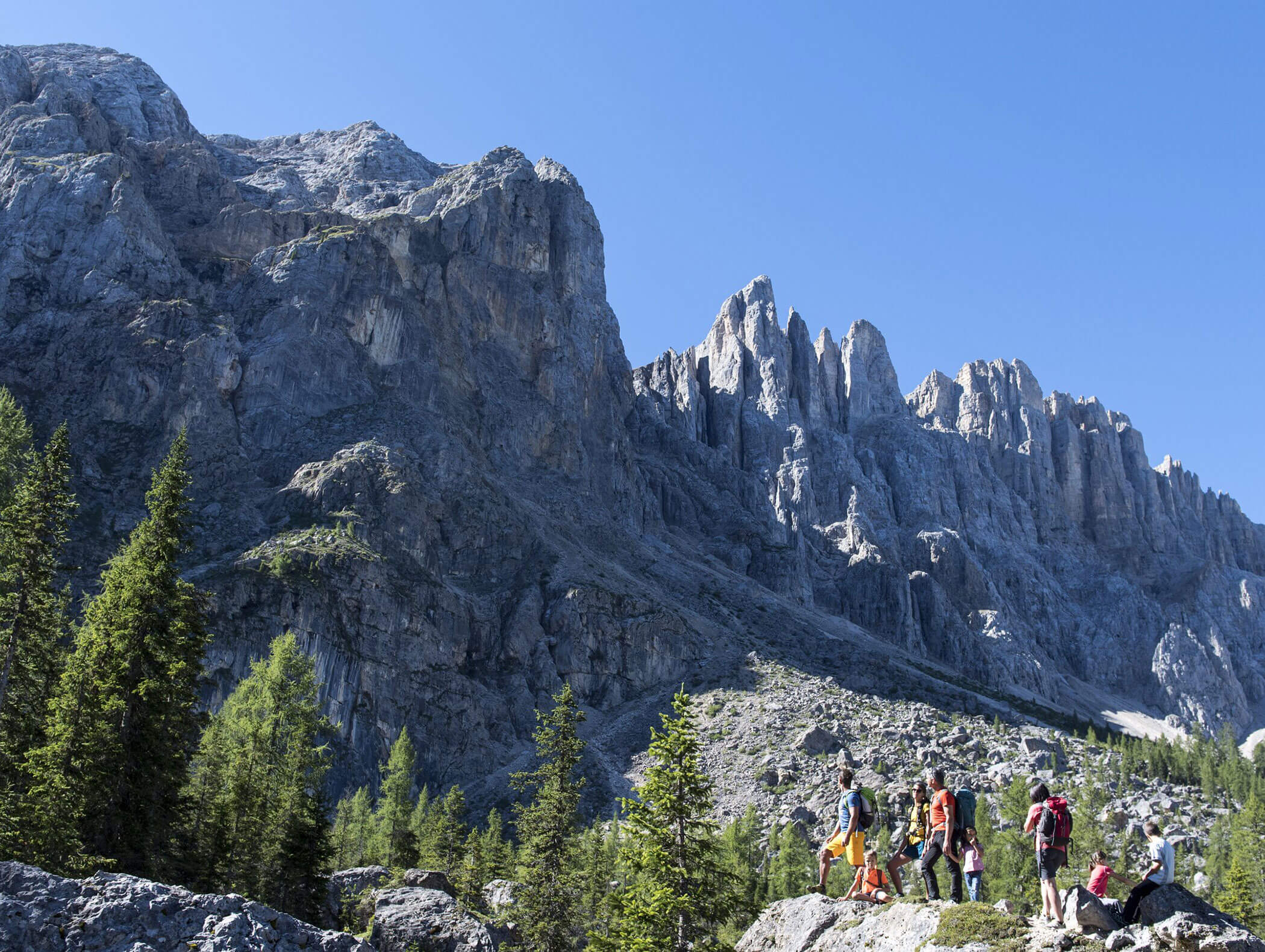 A group of hikers stands on rocky terrain with tall mountains and pine trees in the background. - Hotel Zur Sonne
