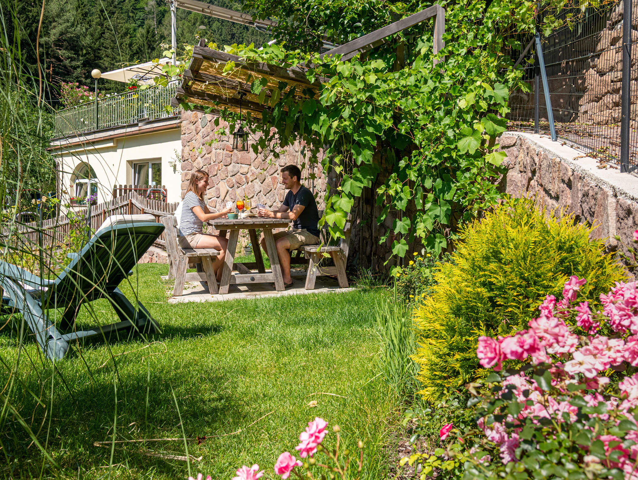 Two people sit at a wooden table in a garden, enjoying drinks surrounded by greenery and pink flowers. - Hotel Zur Sonne