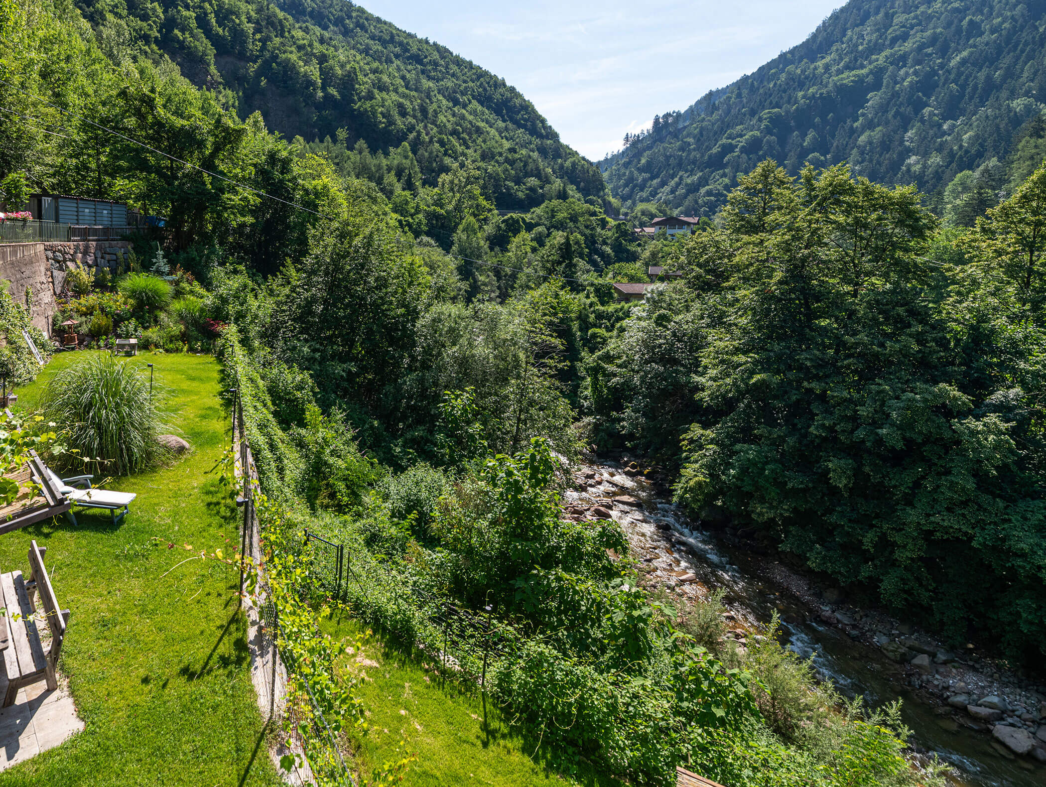 A lush green valley with dense trees, a stream flowing, and a garden with lounge chairs on the left side. - Hotel Zur Sonne