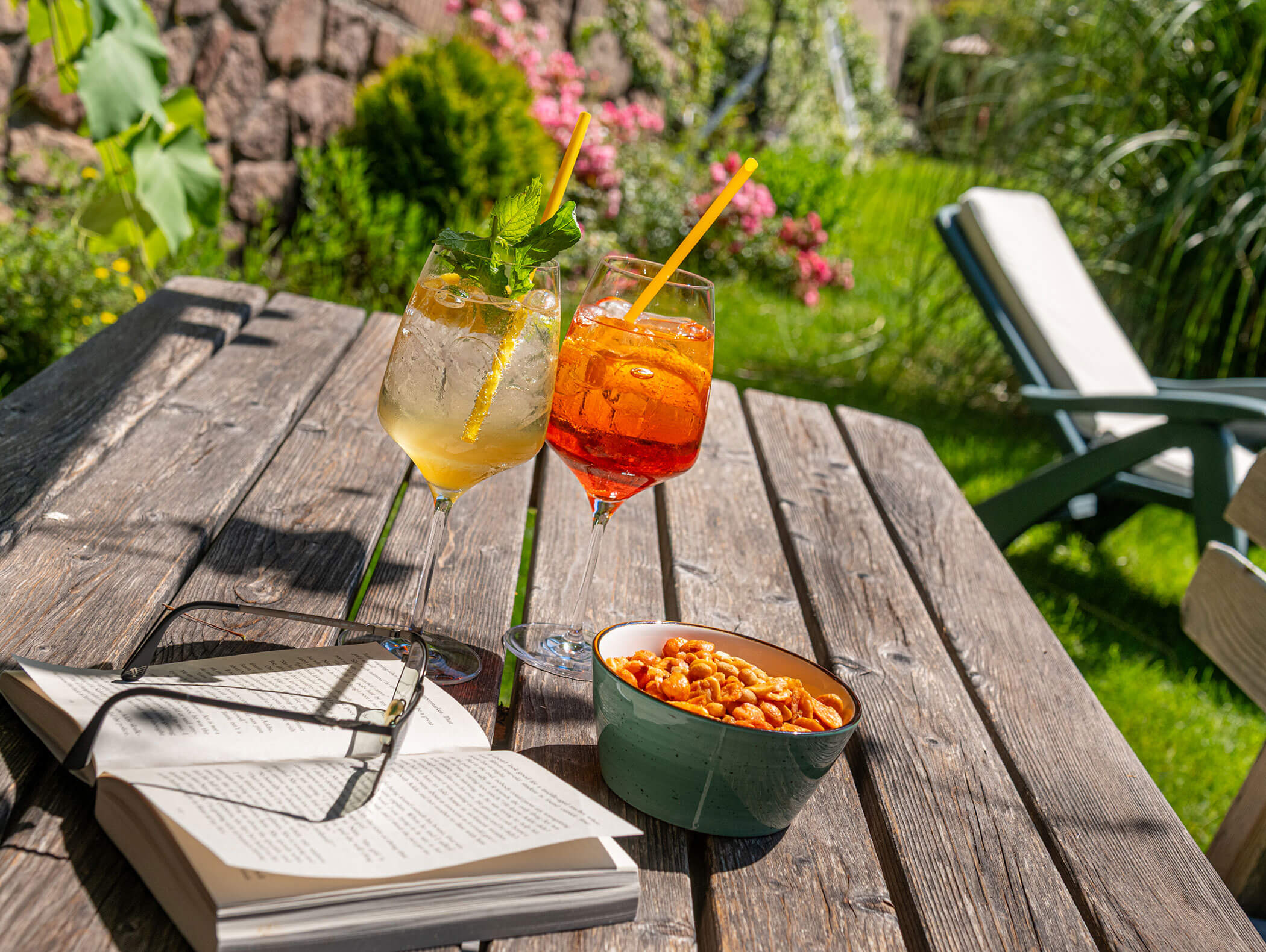 Two cocktails, a bowl of peanuts, an open book, and sunglasses on a wooden table in a sunny garden. - Hotel Zur Sonne