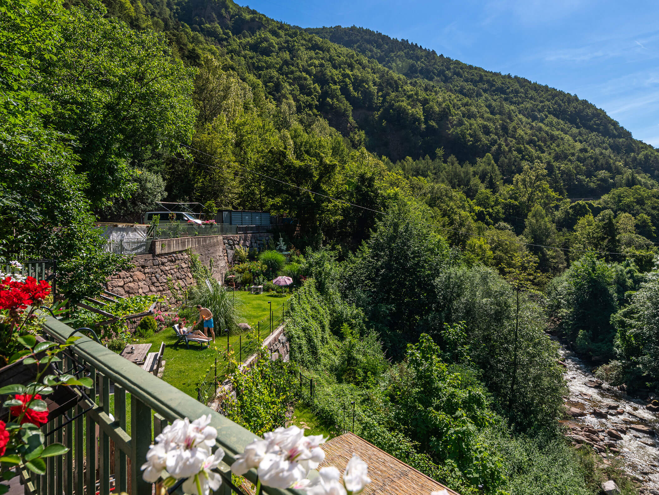 A green mountainside with a river, gardens, and people relaxing near flowers on a sunny day. - Hotel Zur Sonne