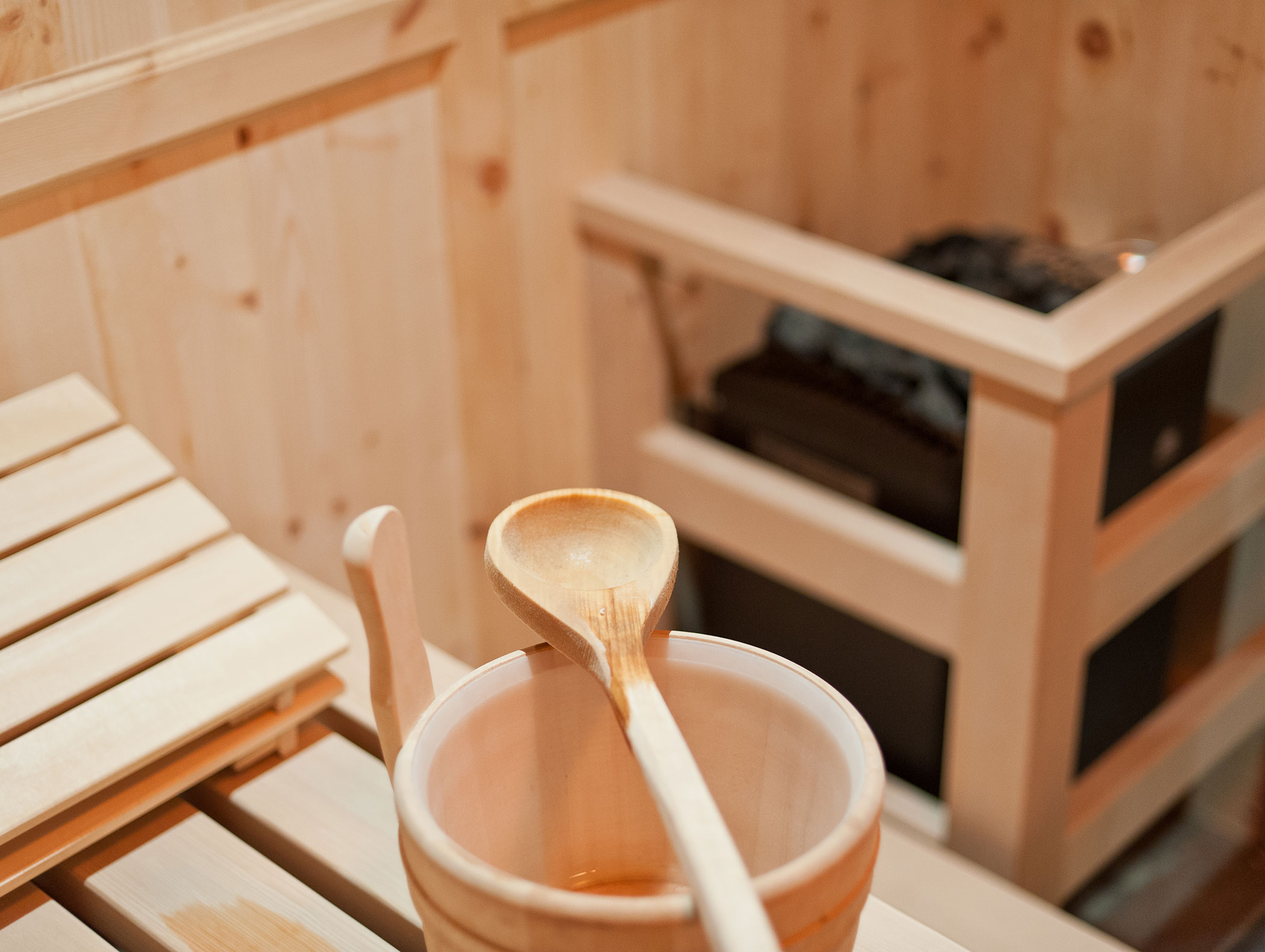 Wooden sauna interior with a bench, bucket, and ladle in the foreground, heater in the background. - Hotel Zur Sonne