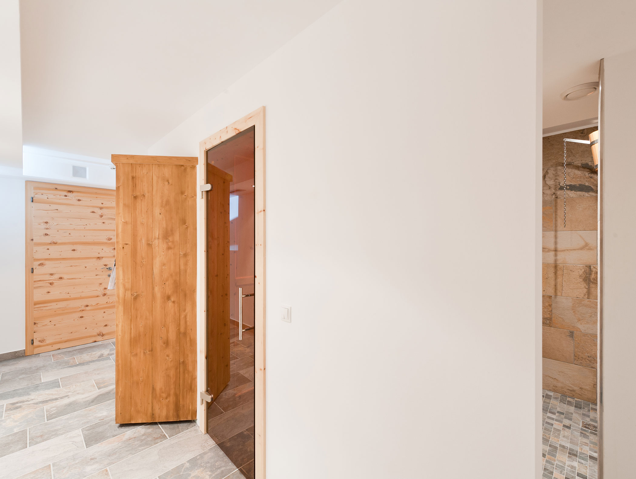 A white hallway with wooden doors, tiled floors, and a glimpse into a shower area with beige stone tiles. - Hotel Zur Sonne