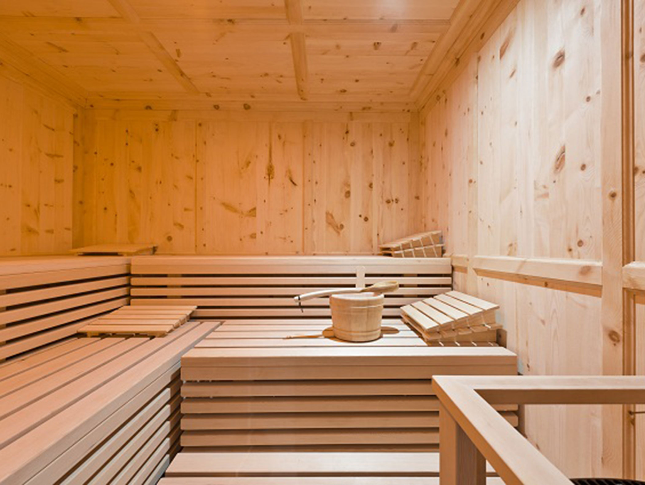 Wooden sauna interior with benches, a bucket, and ladle set on the middle bench against paneled walls. - Hotel Zur Sonne