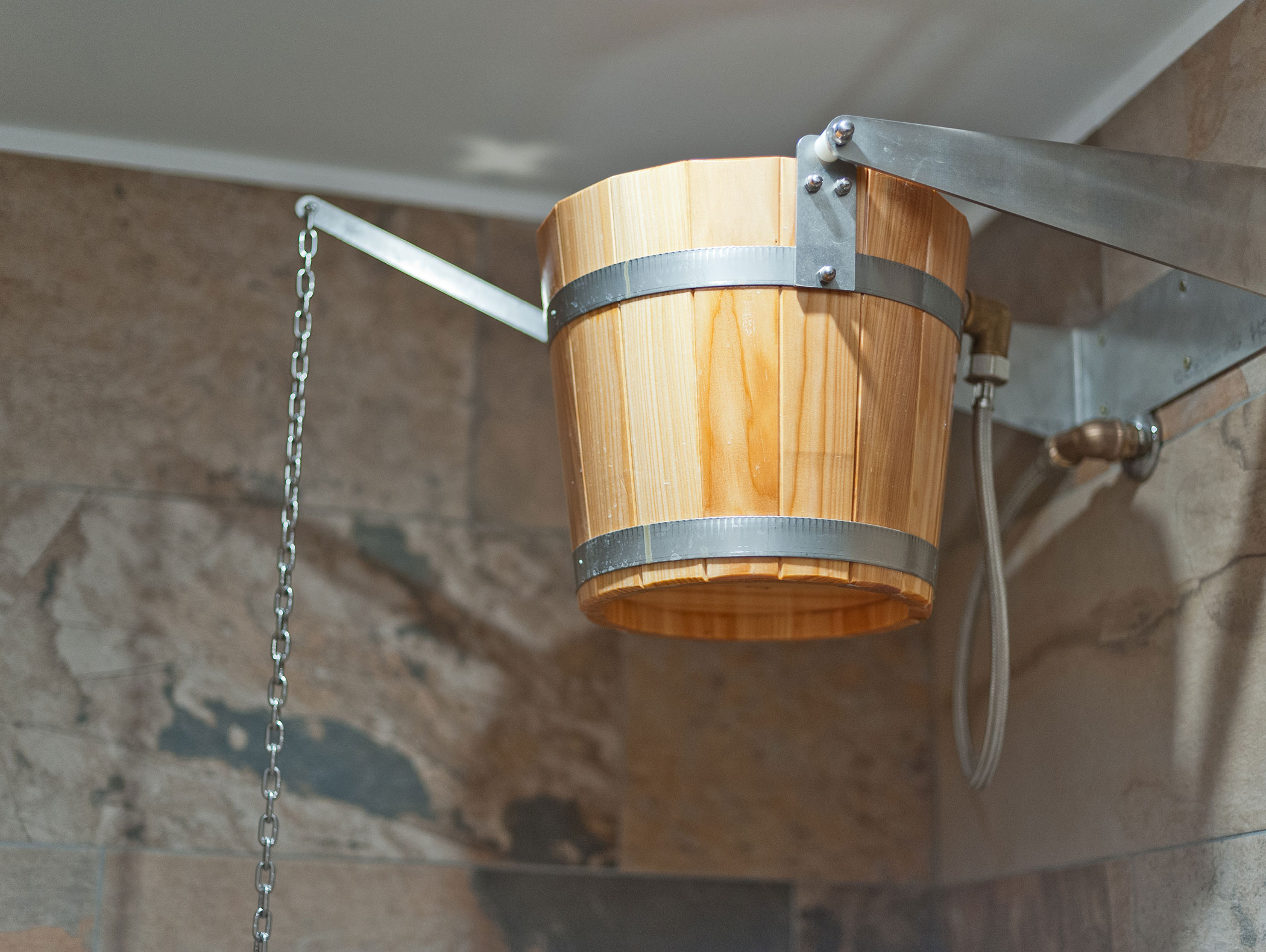 A wooden bucket is mounted on a metal frame in a stone-tiled shower area, with a chain hanging from it. - Hotel Zur Sonne