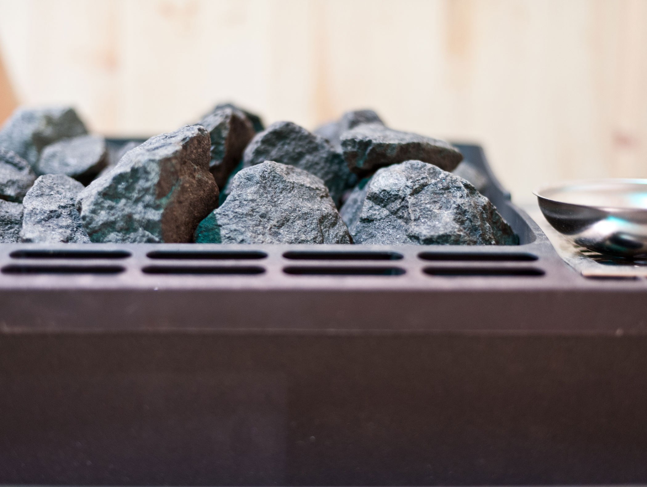 Close-up of dark sauna stones in a metal heater with a blurred wooden wall in the background. - Hotel Zur Sonne