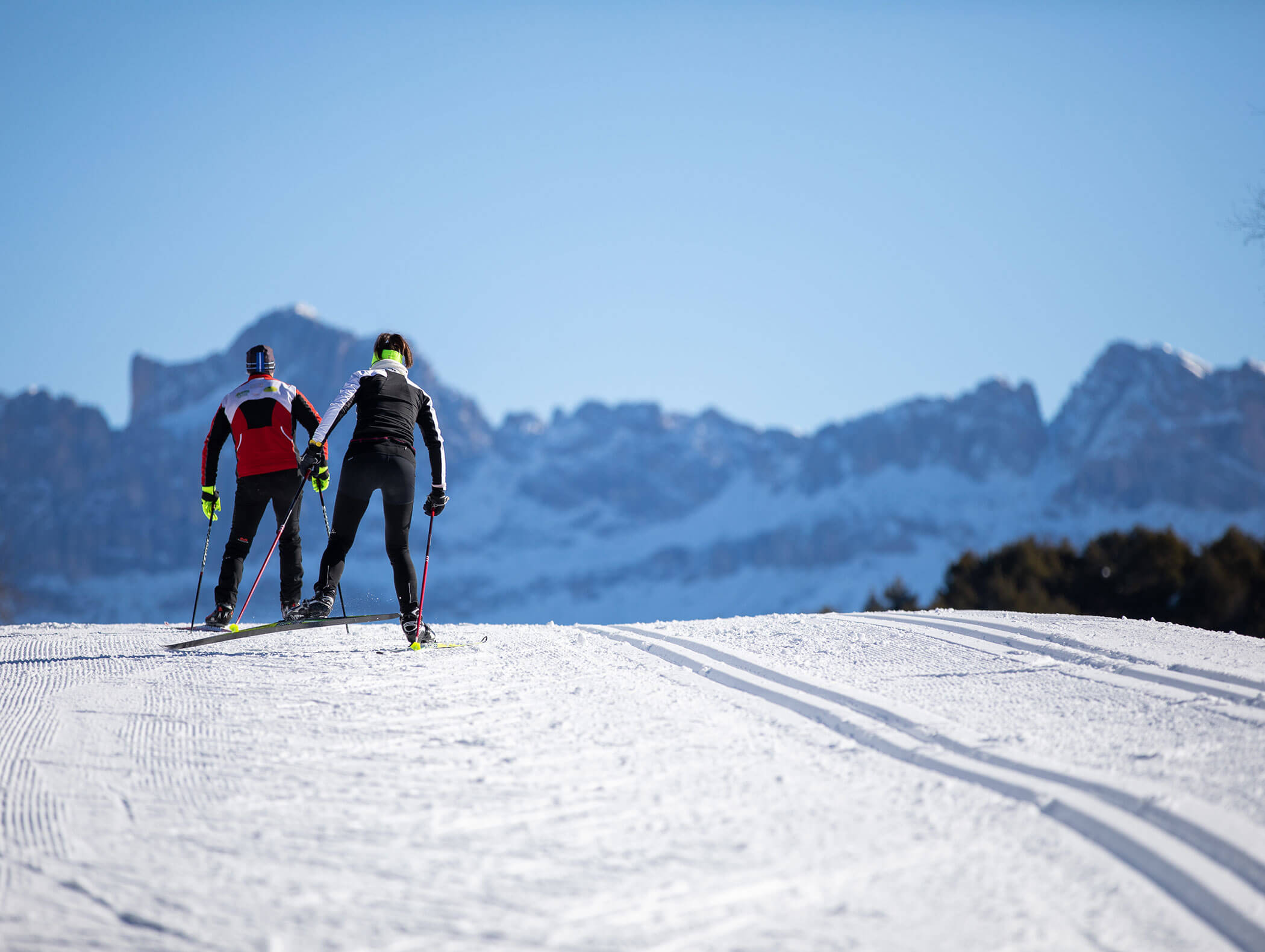Due persone che praticano lo sci di fondo su una pista innevata, con le montagne sullo sfondo e sotto un cielo sereno. - Hotel Zur Sonne