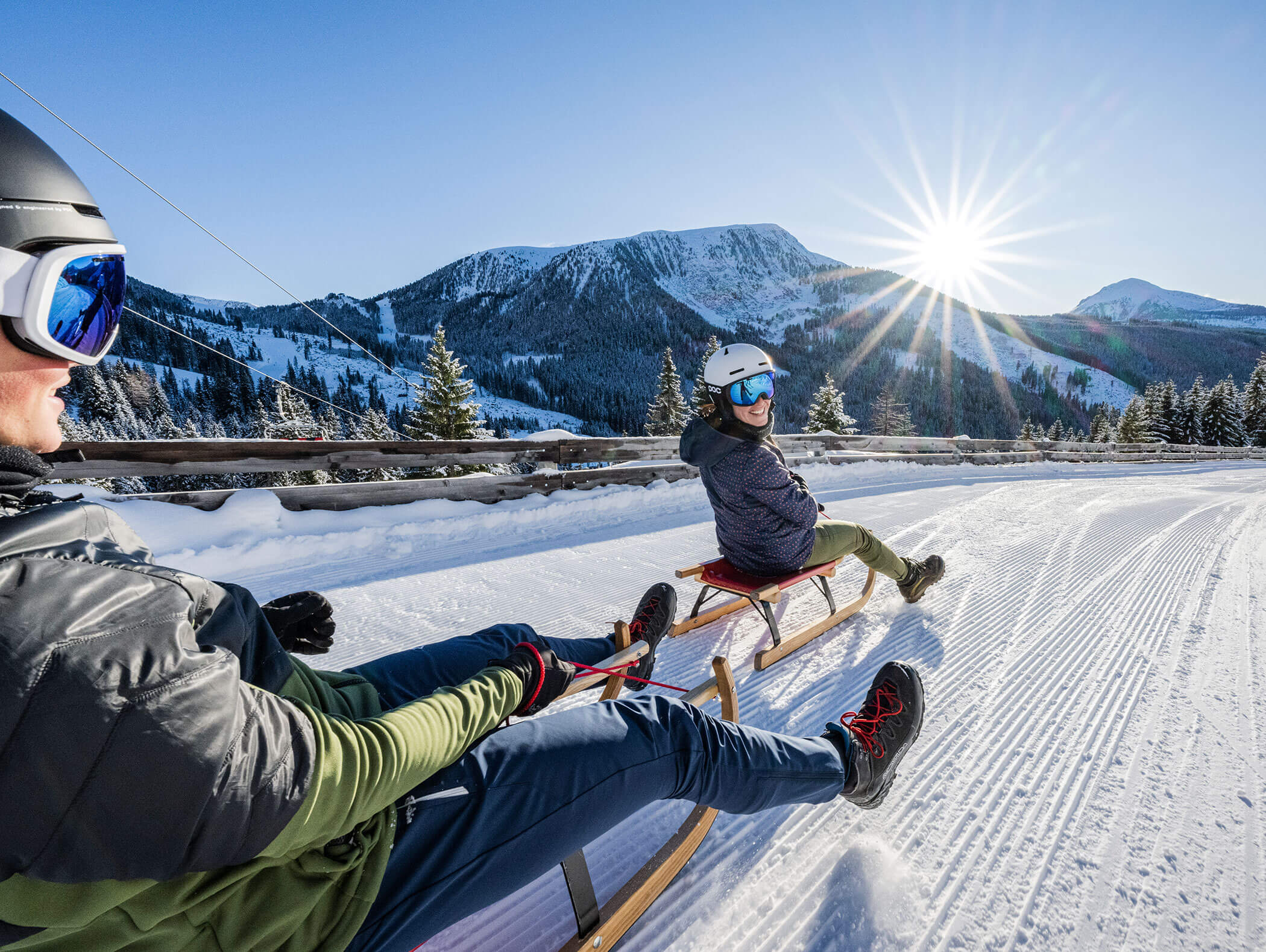 Due persone che scendono in slitta lungo un pendio innevato sotto un sole splendente, con pini e cime di montagna sullo sfondo. - Hotel Zur Sonne