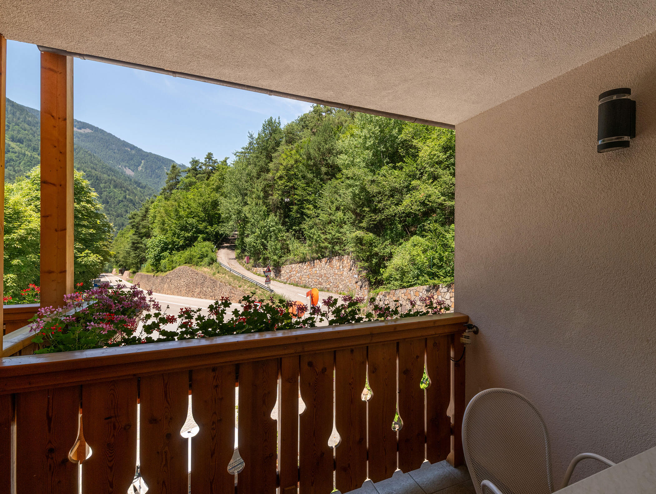 Vista da un balcone con ringhiera in legno, fiori e sedie, affacciato su una strada tortuosa e alberi verdeggianti. - Hotel Zur Sonne
